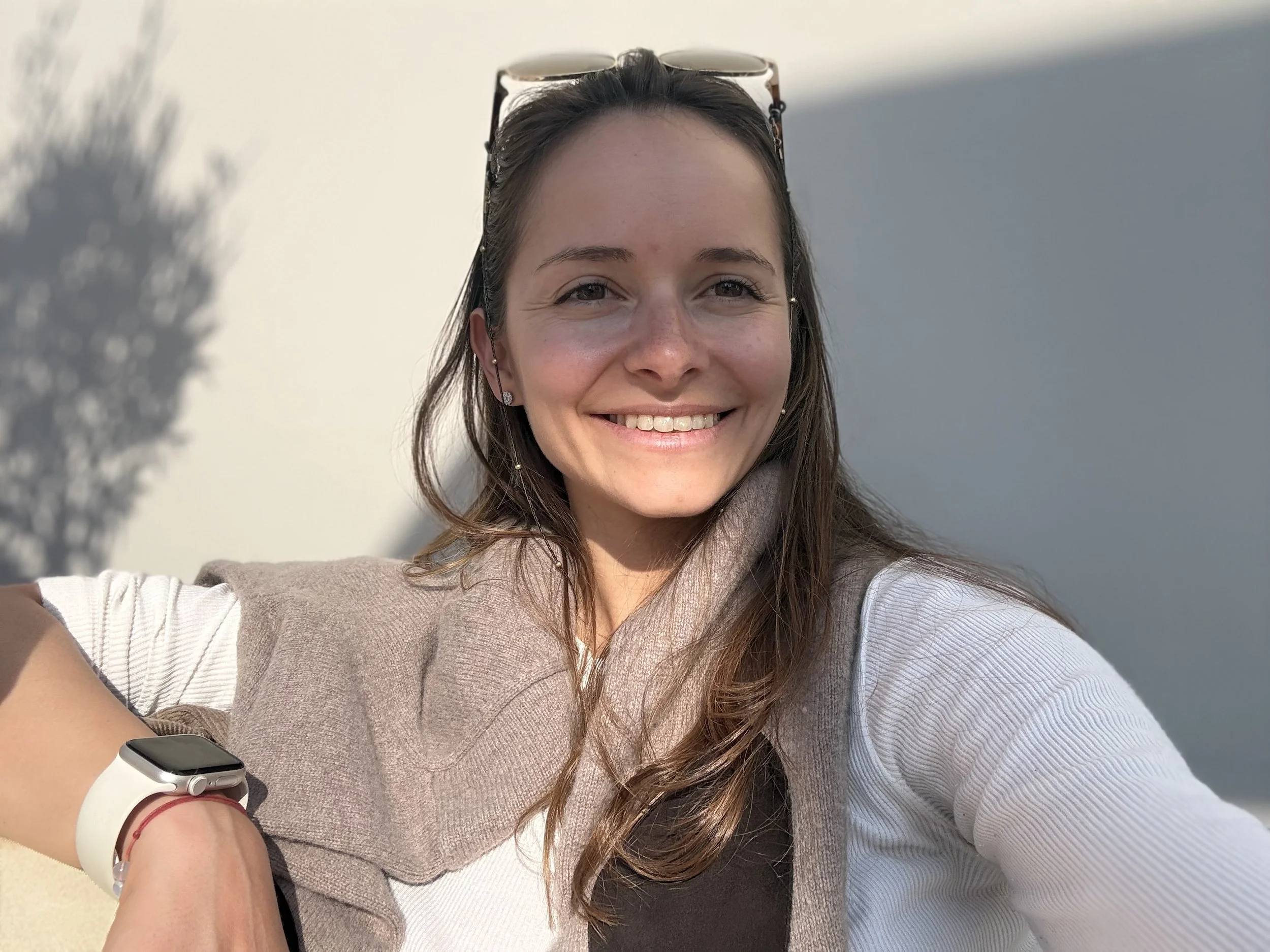 A young woman smiling, wearing sunglasses on her head and a smartwatch on her wrist, with sunlight and a shadow of a tree in the background.