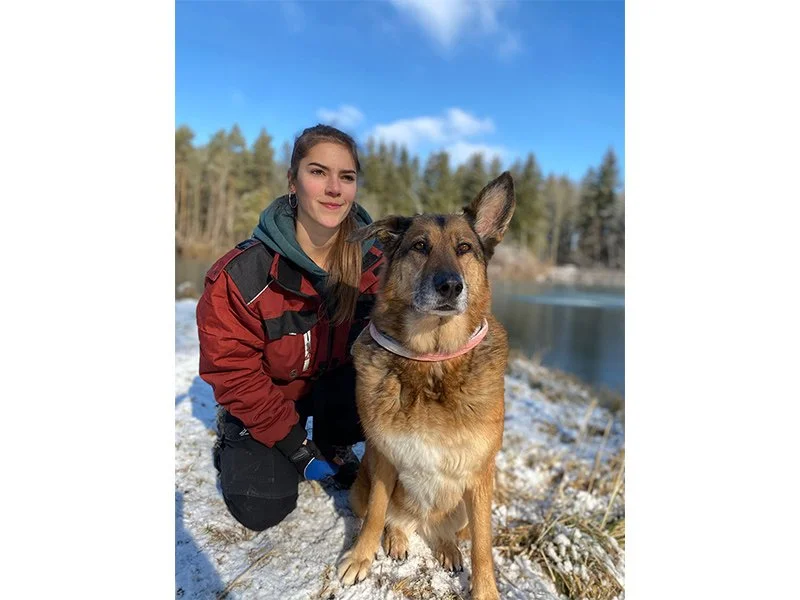 Ein junges Mädchen mit braunen Haaren und einem roten Jacke kniet neben einem großen braunen Hund mit schwarzen Ohren am Ufer eines Sees im Winter mit Schneeboden und Baum im Hintergrund, blauer Himmel mit Wolken.