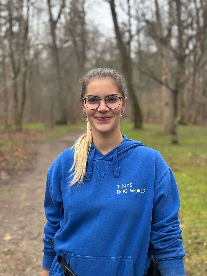 Junge Frau im blauen Hoodie mit Text "TINI'S DOG WORLD" steht im Park