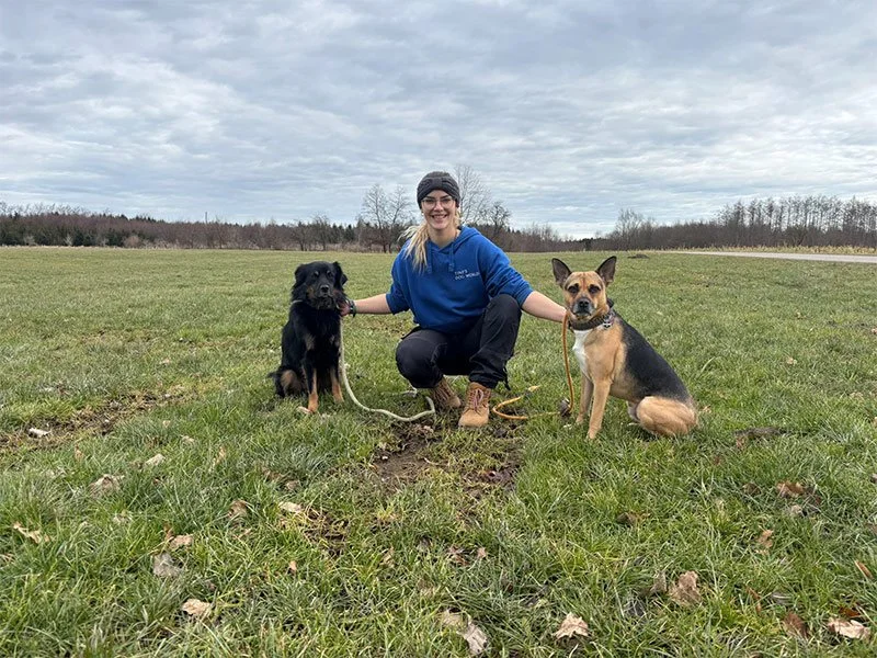 Eine lachende Frau in blauer Kapuzenjacke mit zwei Hunden auf einer Wiese im Freien.