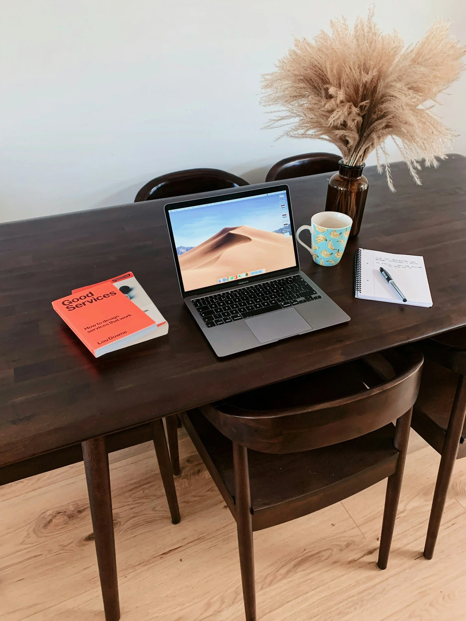 A dark wooden dining table with a closed MacBook, a stack of two books titled 'Good Services' and 'How to Design Services,' a notebook with a pen, a ceramic mug with bananas, and a large brown vase with beige pampas grass.