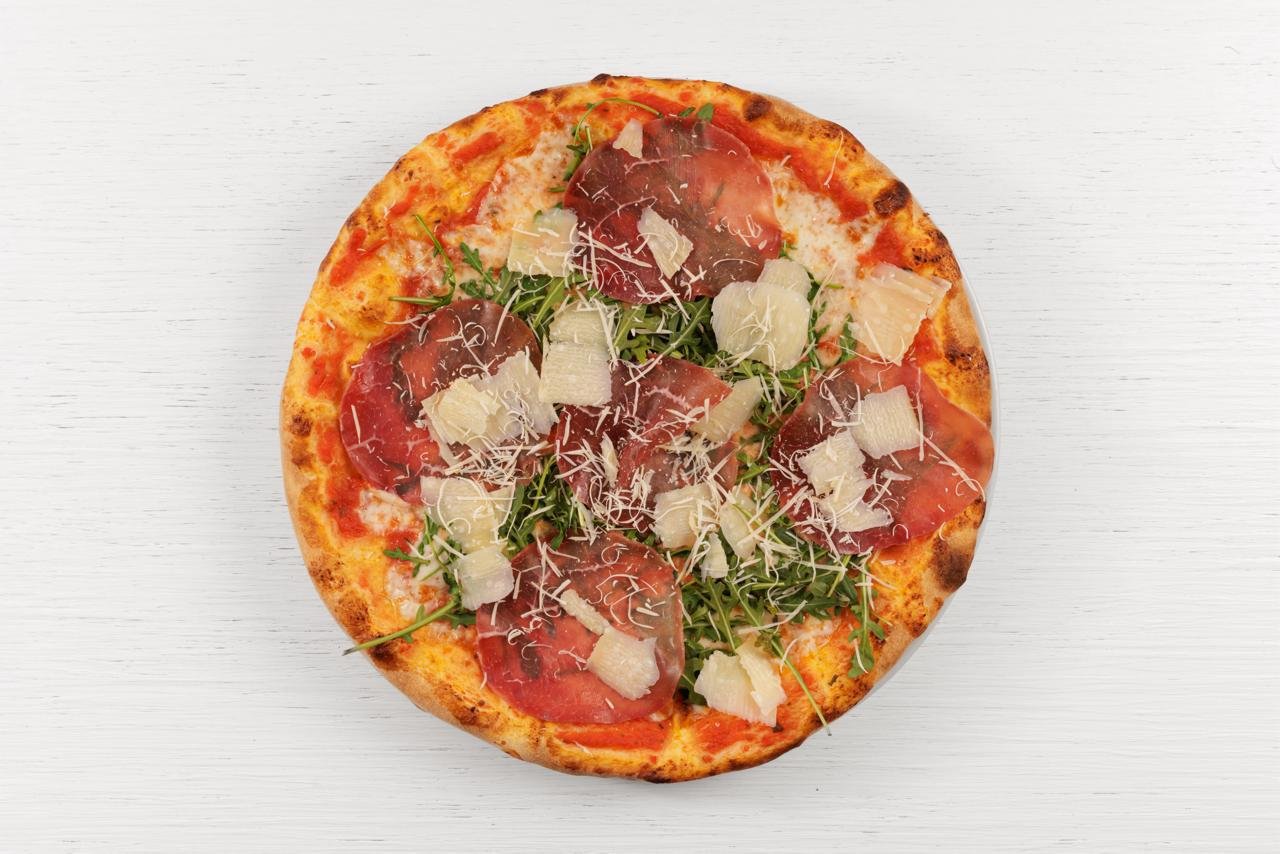 Pizza con pomodoro, prosciutto, rucola e scaglie di formaggio su superficie bianca.