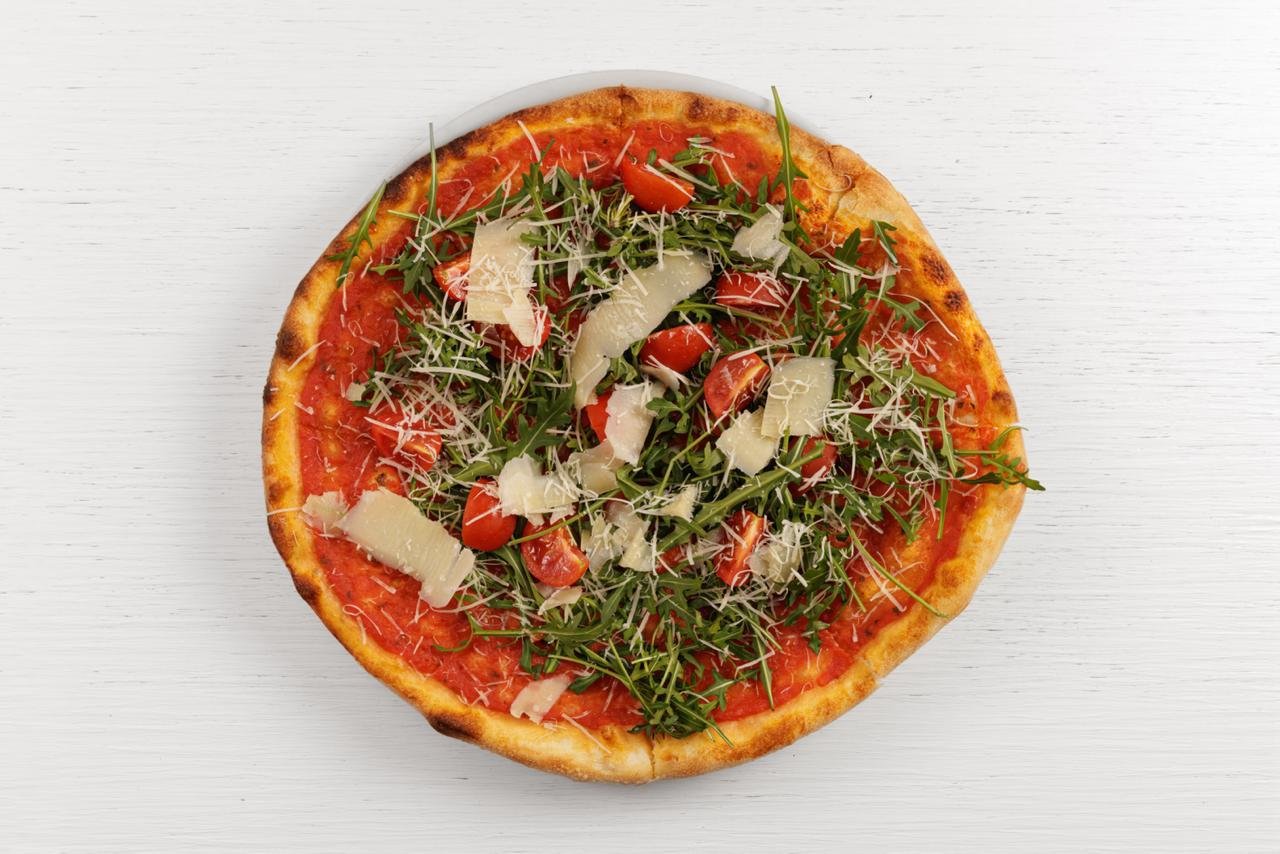 Fetta di pizza con pomodorini, rucola e scaglie di formaggio su un tavolo di legno bianco.
