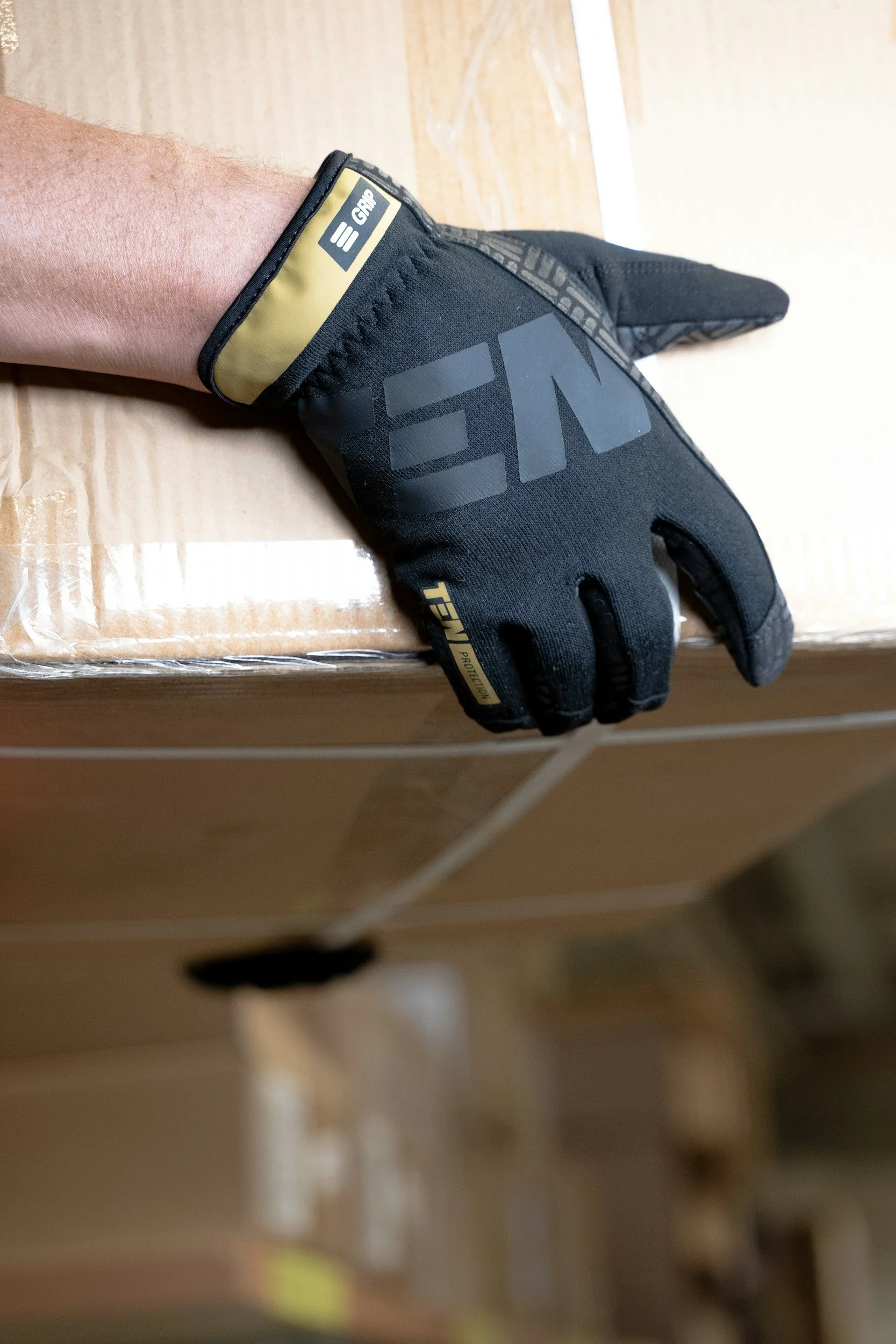A person's hand resting on a cardboard box, wearing a black and yellow work glove with branding.