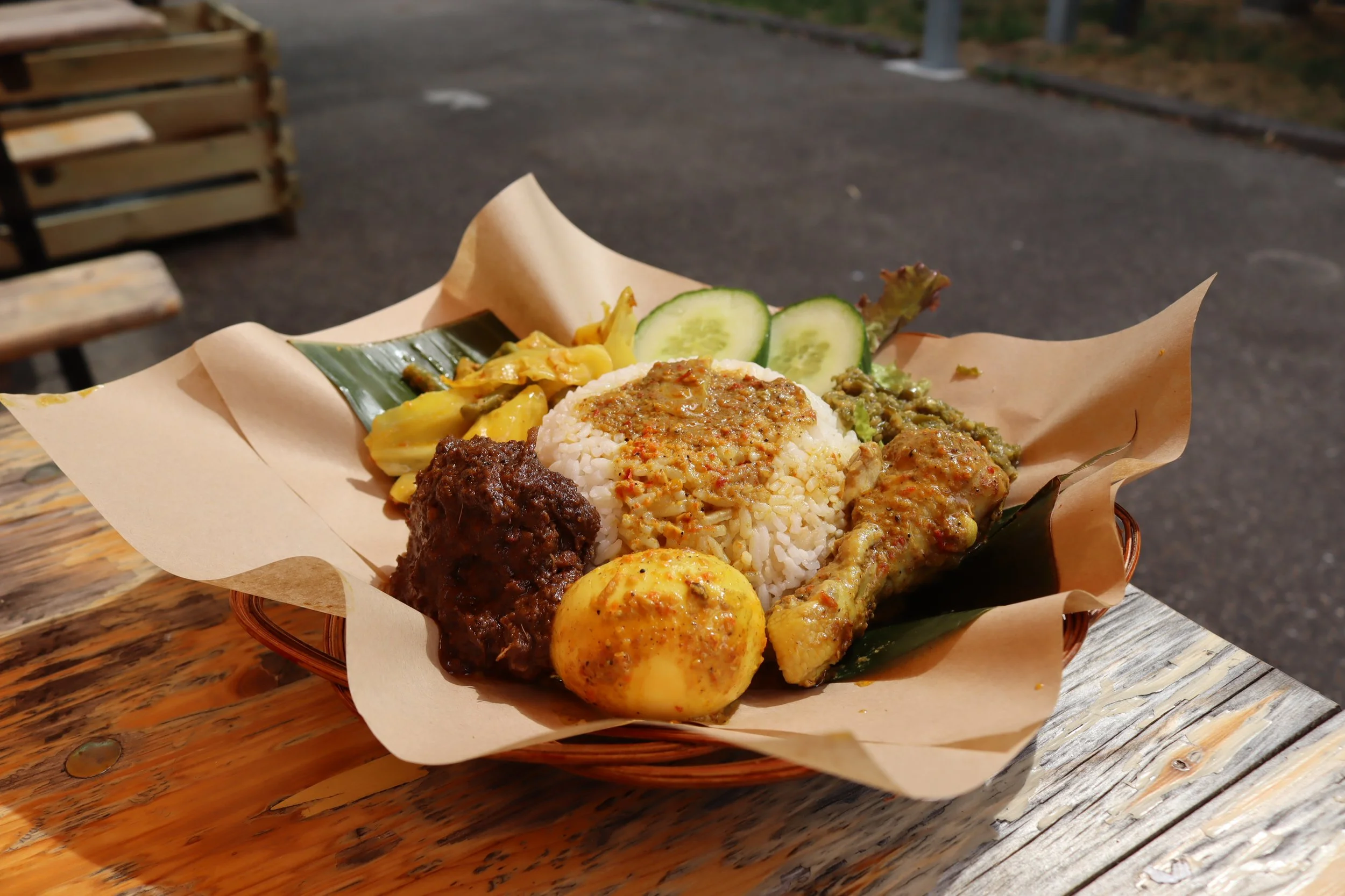 A serving of Indonesian food with rice, vegetables, chicken, and an egg, arranged in a basket with paper liner outside on a wooden table.