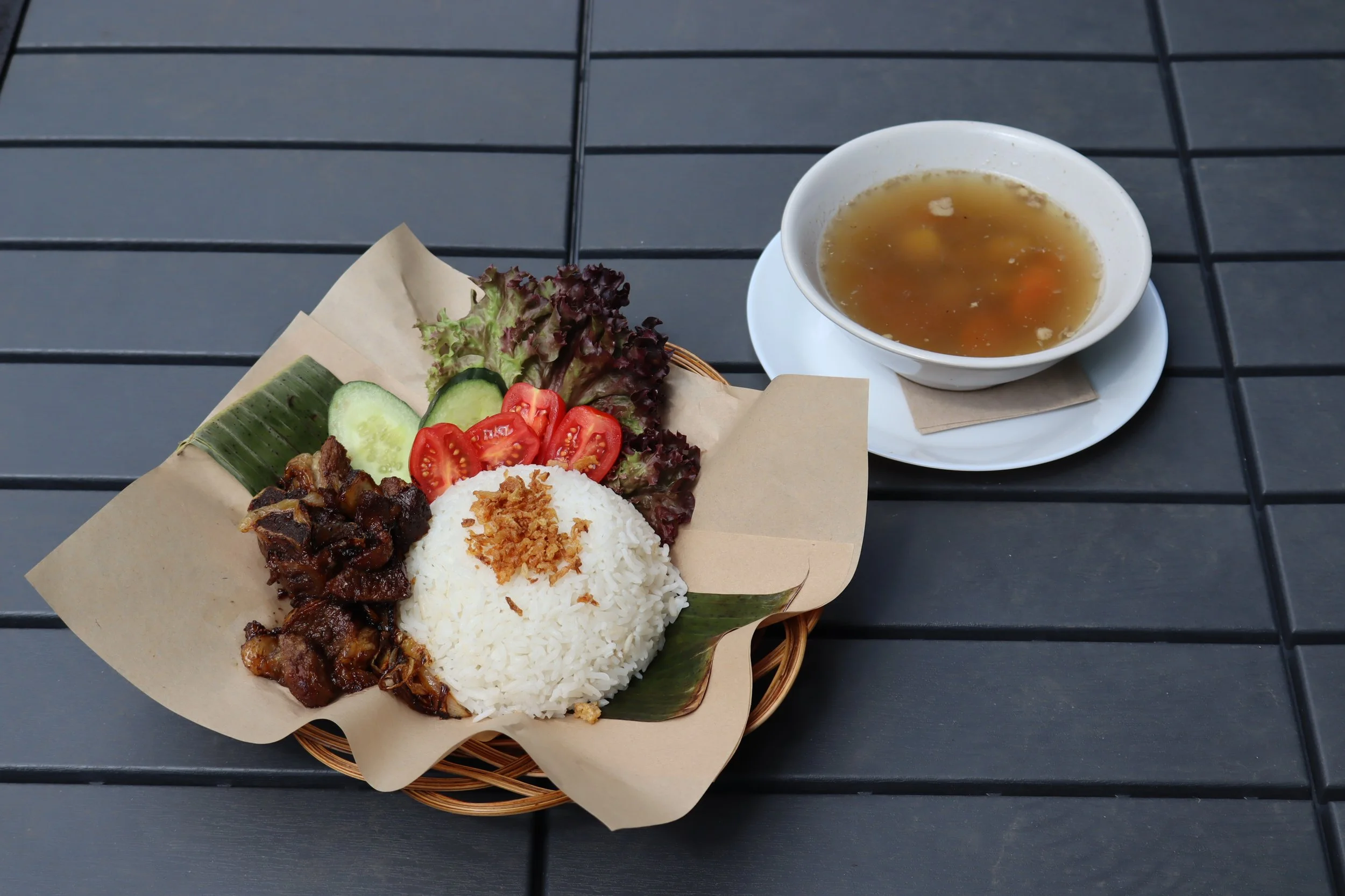 Plate of rice with fried chicken served with fresh vegetables and salad, accompanied by a bowl of clear soup with vegetables on a black table.