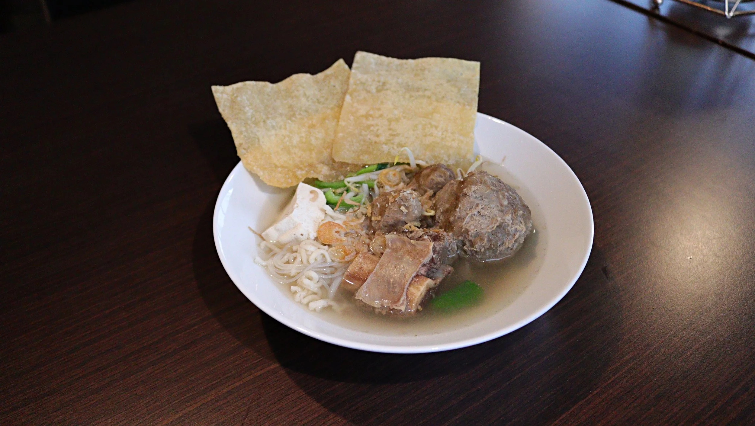 A white bowl containing noodle soup with beef meatballs, vegetables, and crispy crackers, placed on a dark wooden table.