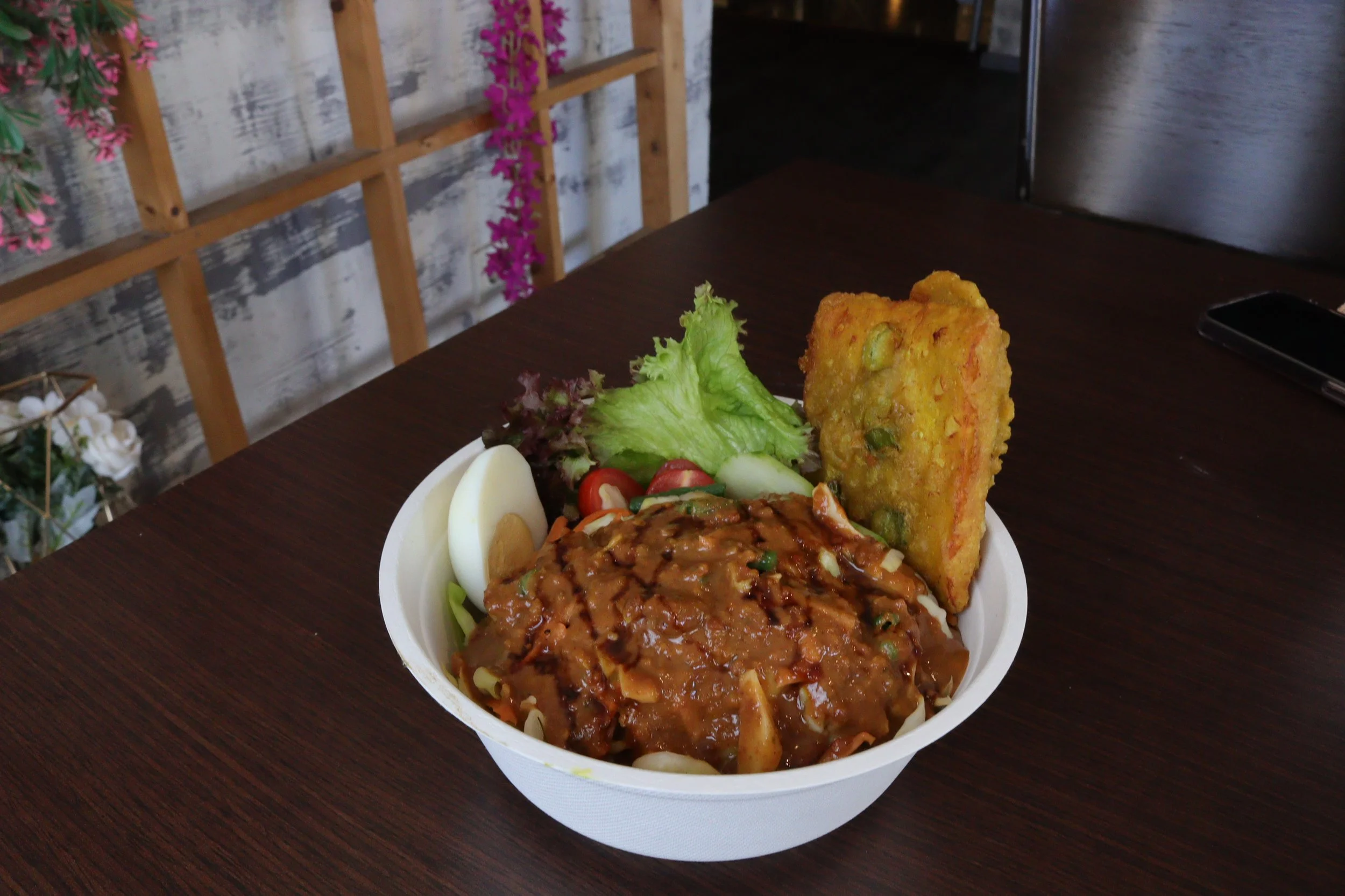 A white bowl of Indian-style curry topped with gravy, served with a boiled egg, lettuce, cherry tomatoes, cucumbers, and a large piece of vegetable pakora on the side.