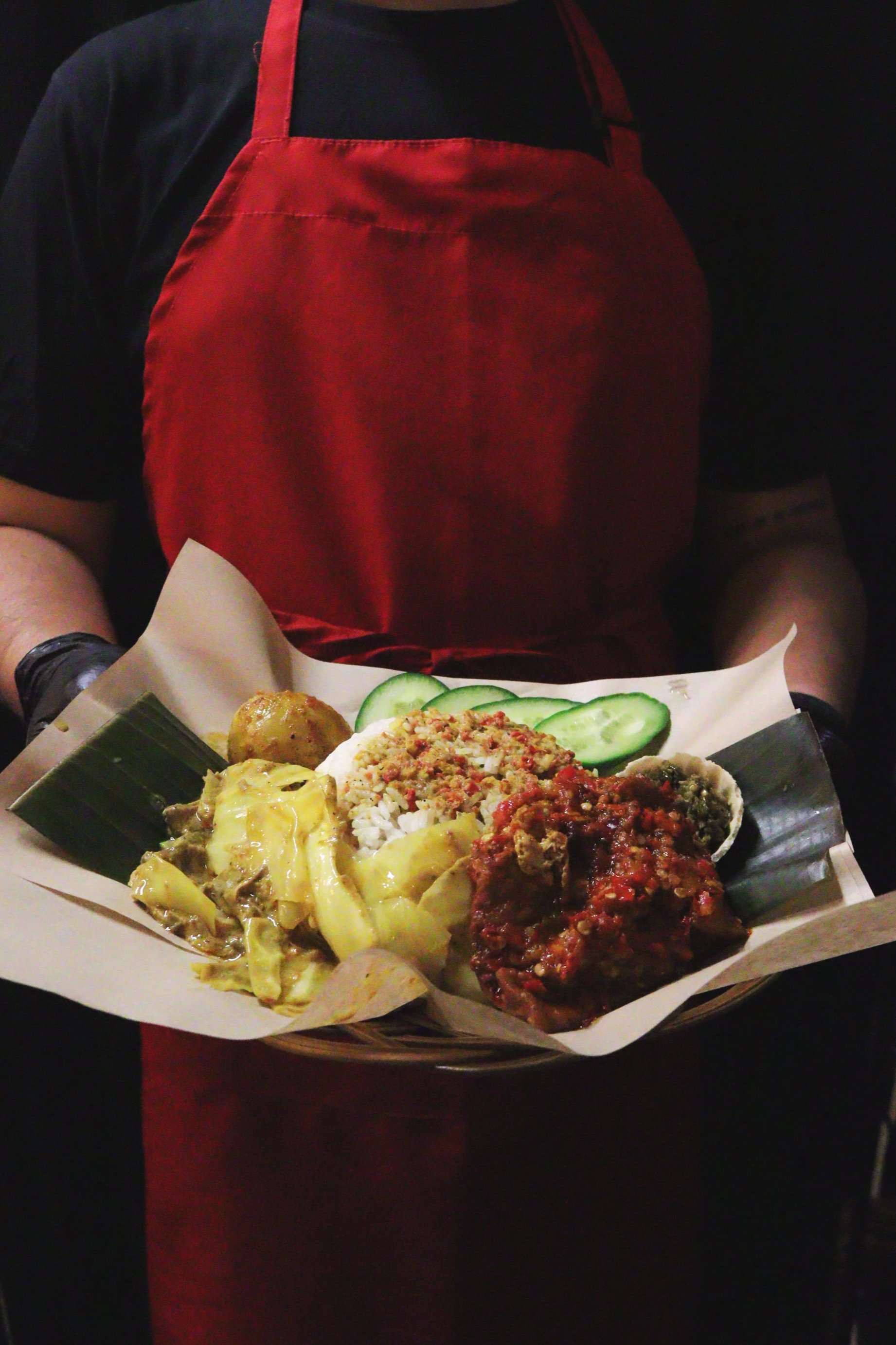 Person in black shirt and red apron holding a tray of mixed Asian dishes, including rice, cucumber slices, and various curries.