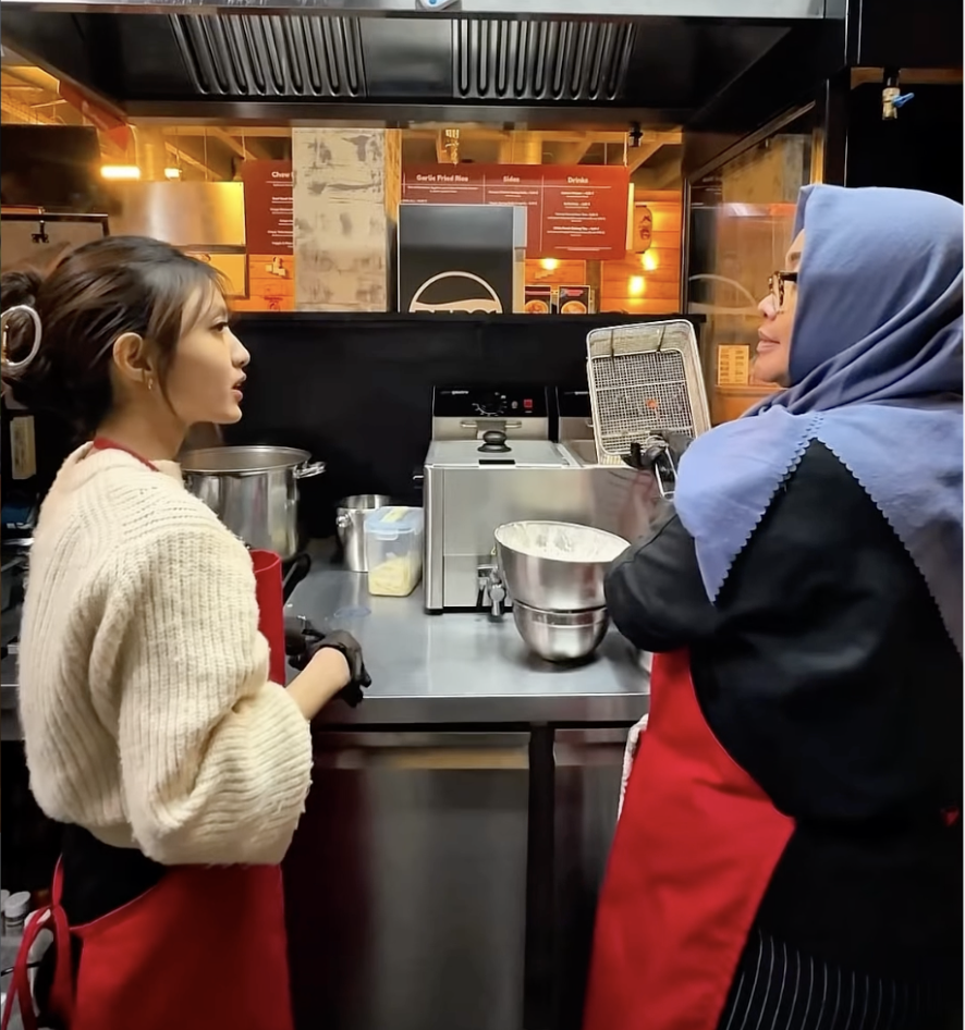 Two women in a kitchen—one young woman with an updo hairstyle and a beige sweater, and an older woman wearing a blue headscarf and glasses—stand facing each other near a countertop with cooking appliances, engaging in conversation.