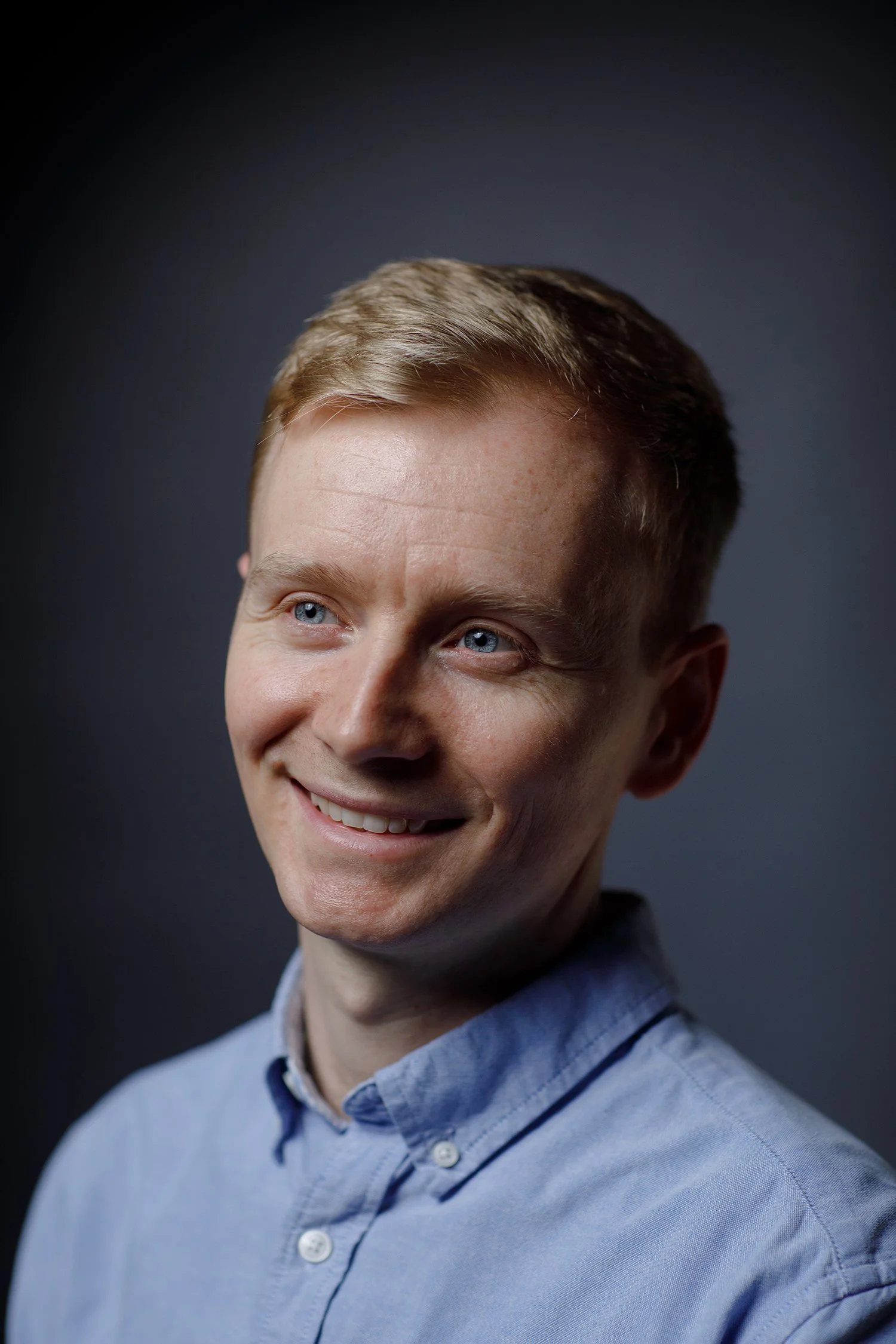 Andrew Wilson has blonde hair and blue eyes, is smiling and looking to his left. He is wearing a light blue collared shirt against a dark background.