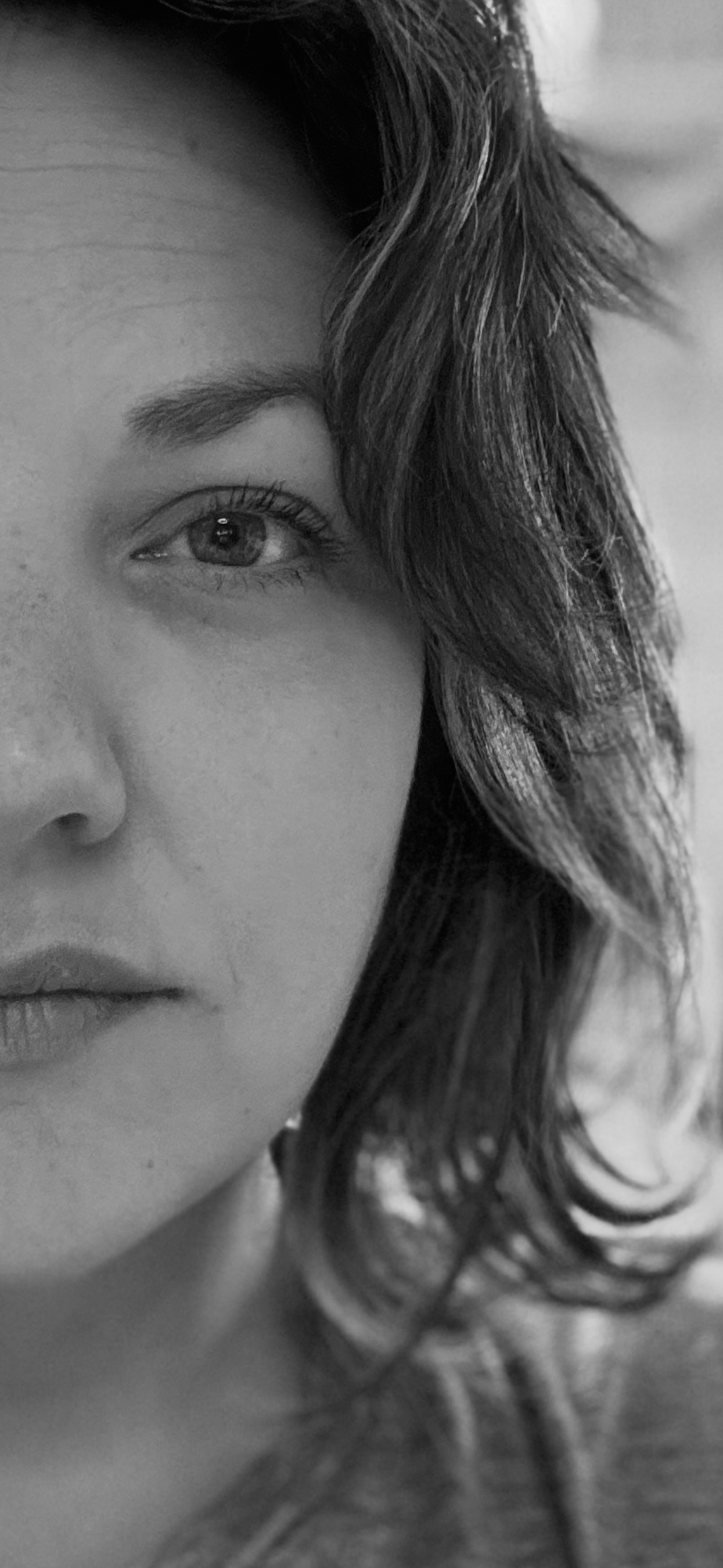 Nahaufnahme eines Gesichts mit sichtbarem Auge, einer dichten Augenbraue, einem Teil der Nase und Lippen; dunkelgelocktes Haar, das teilweise das Gesicht bedeckt; Schwarz-Weiß-Fotografie.