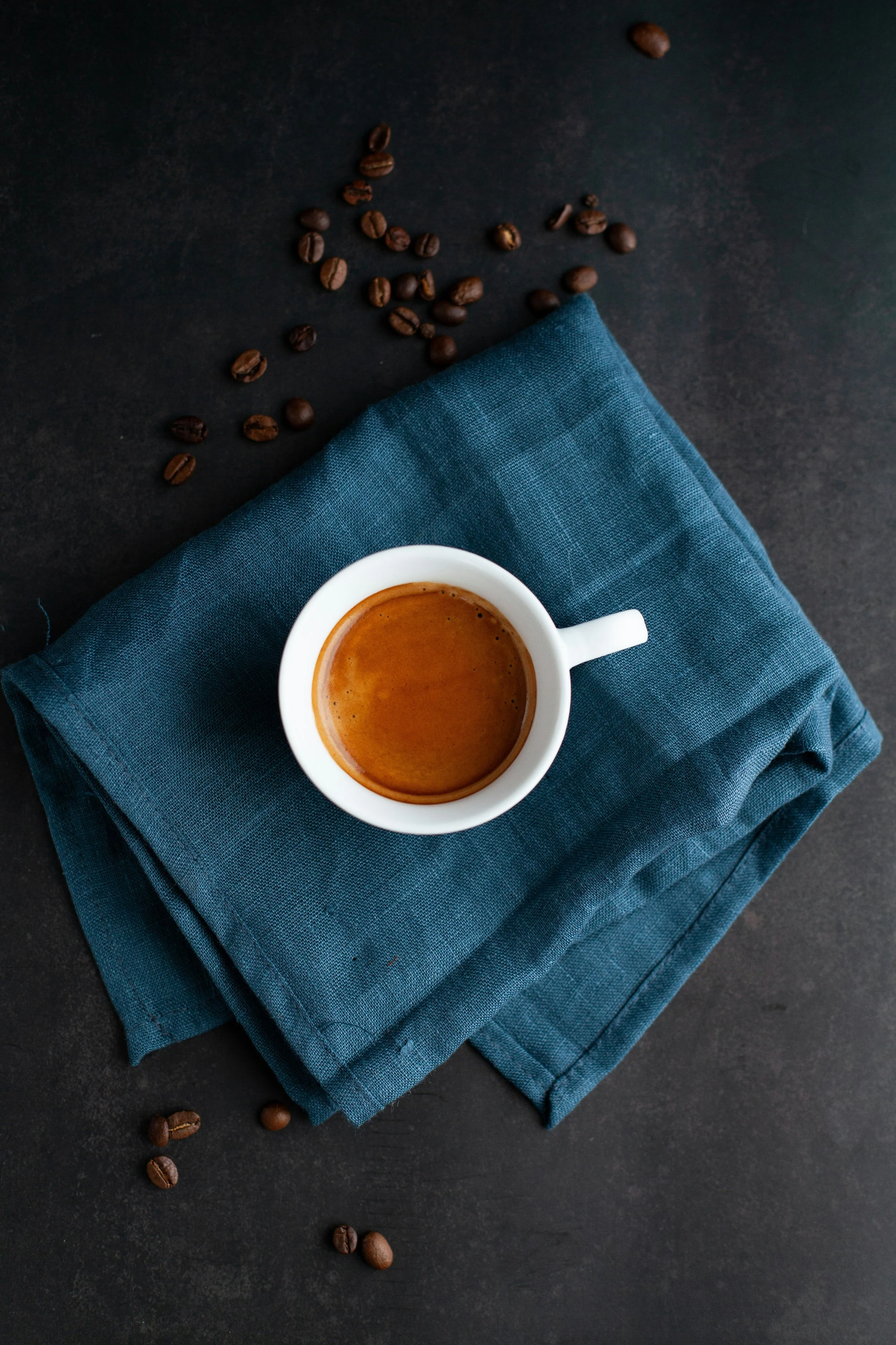 A white coffee cup filled with espresso on a dark surface, resting on a folded blue cloth napkin with scattered coffee beans around.