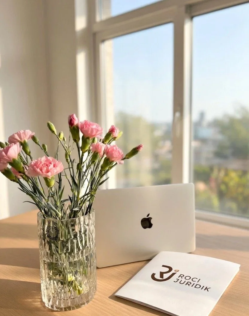 En bukett pinka blommor i en glaskanna, ett stängt vit äpple, ett öppet manussblad med texten "Roci Juridik", bredvid varandra på ett träbord nära ett stort fönster.