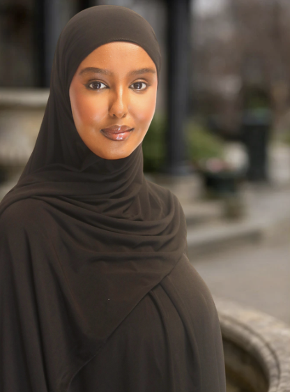 En kvinna i svart hijab står utomhus med en stadsmiljö i bakgrunden.