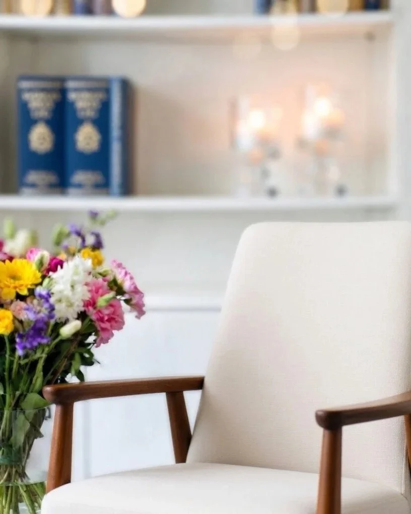 En vit fåtölj med träarmstöd bredvid en bukett av färgglada blommor, i ett svalt inomhusrum.