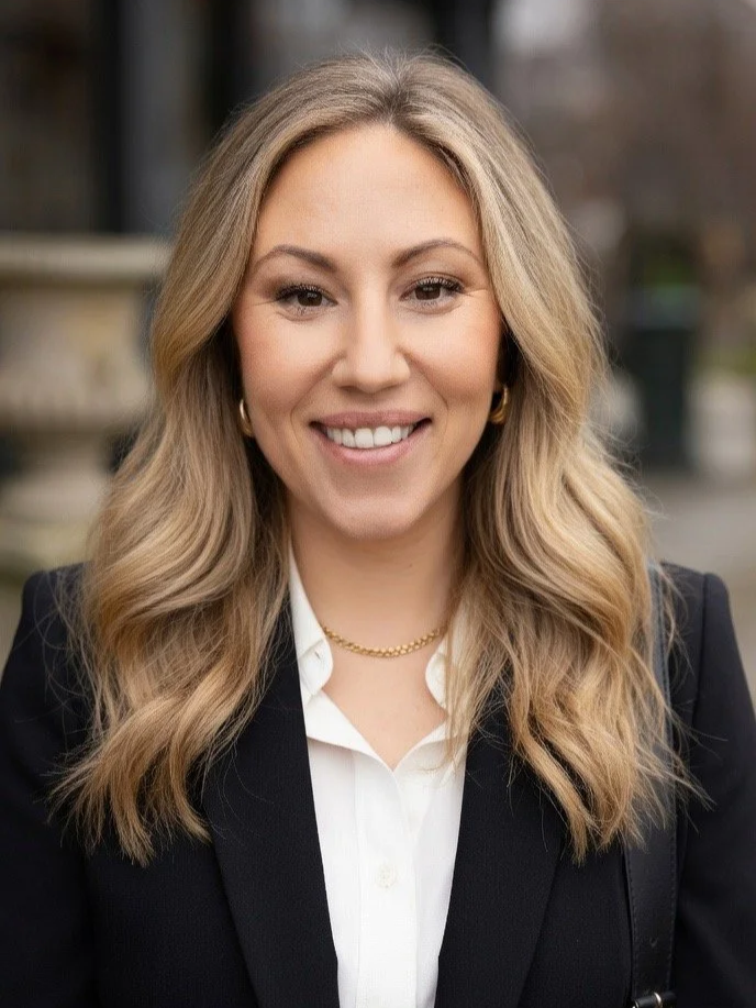 En kvinna med blont, lockigt hår som ler, bär en vit skjorta, svart blazer och guldkedja, utomhus.