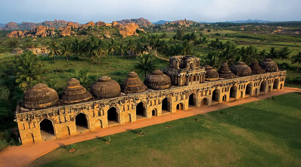 A historic stone building with multiple domed structures on top, set in a lush green landscape with palm trees, rocky hills in the background, and a cloudy sky.