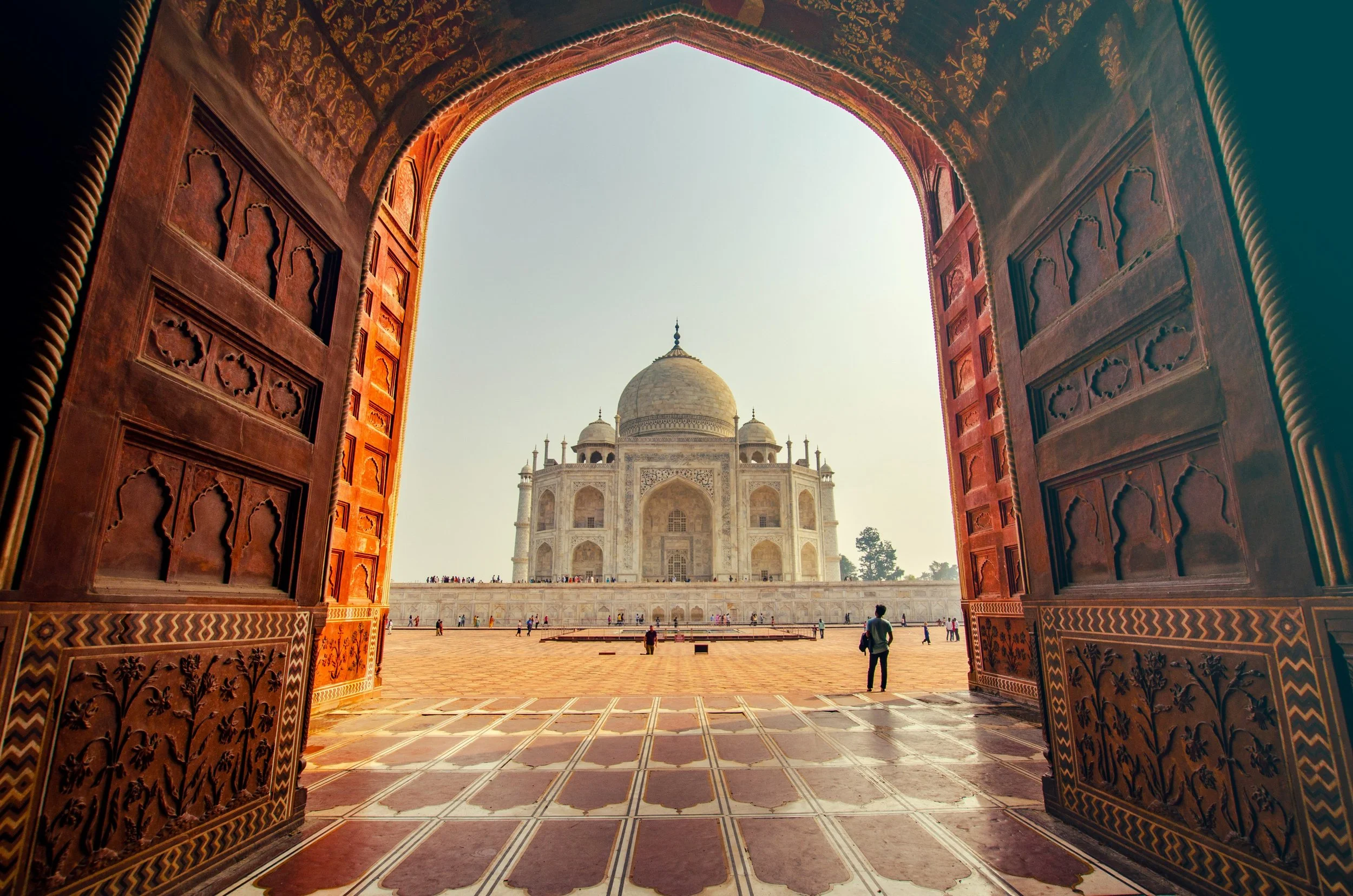 View of the Taj Mahal seen through ornately decorated wooden gates, with the monument's white marble dome and minarets visible in the distance and visitors walking around.