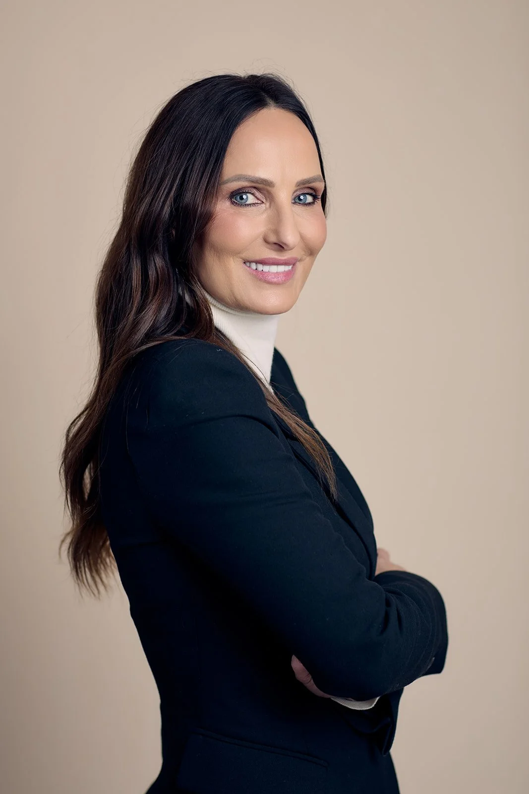 A woman with long dark hair, blue eyes, and fair skin smiling, dressed in a black blazer over a white turtleneck, standing with arms crossed against a beige background.