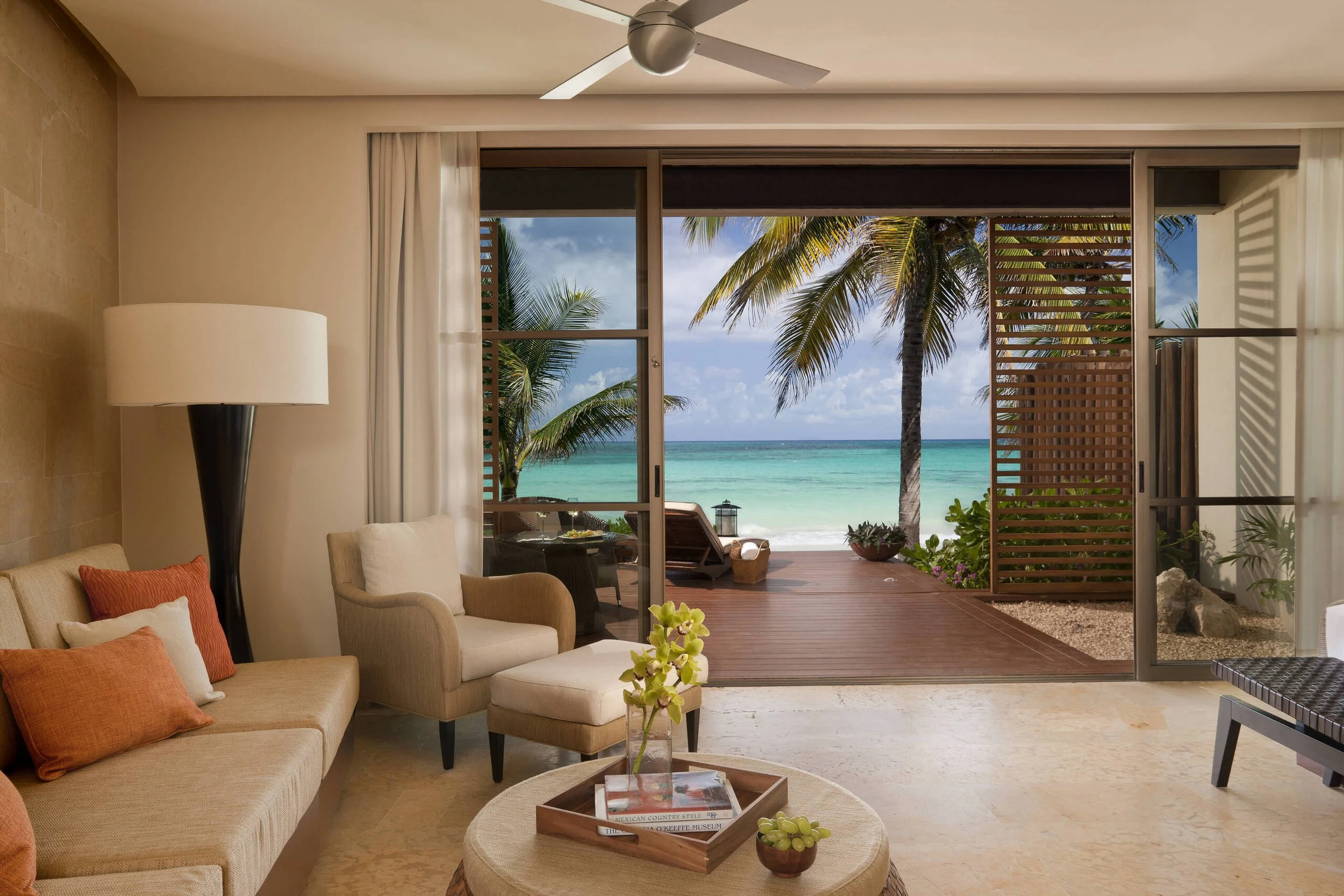 Living room with beige sofa and armchair, colorful pillows, wooden coffee table with books and plants, sliding glass door open to a balcony with ocean view and palm trees