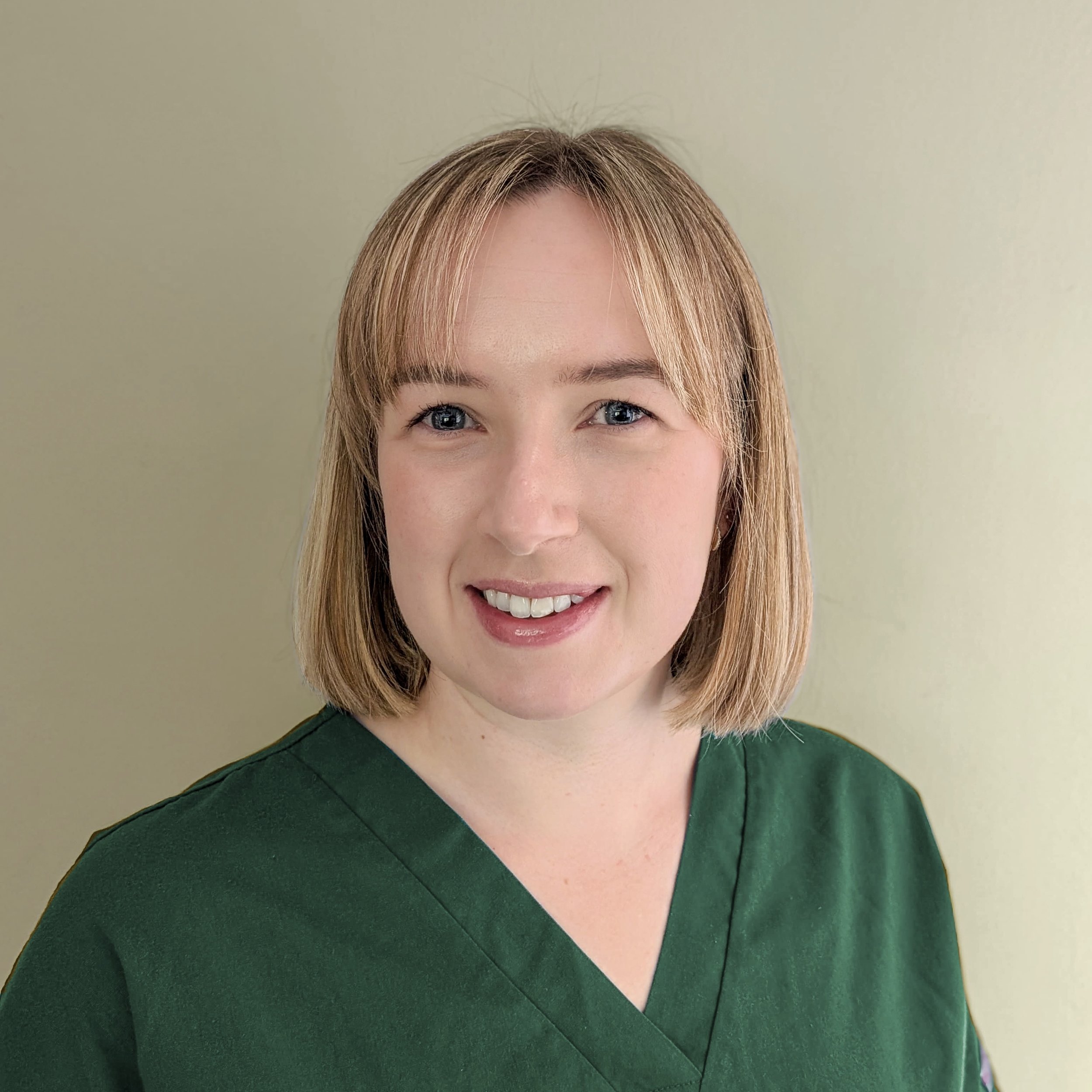 A woman with short blonde hair, wearing a dark green top, smiling against a light-colored wall.