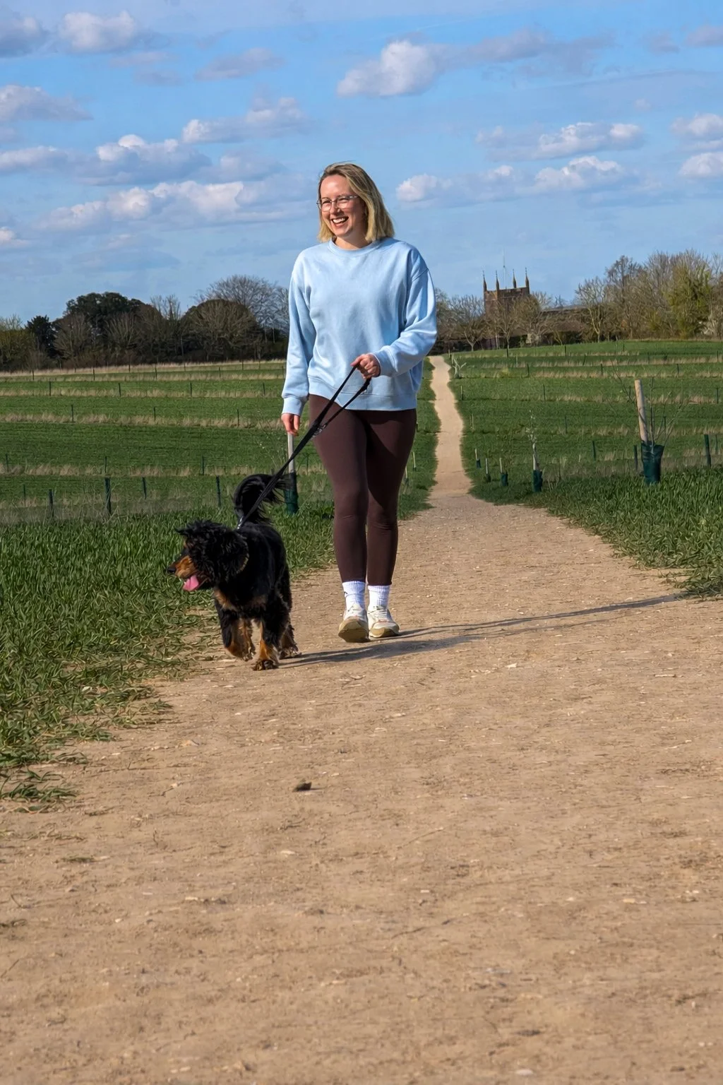 A woman walking a black and tan dog on a dirt path through a green field under a partly cloudy sky.