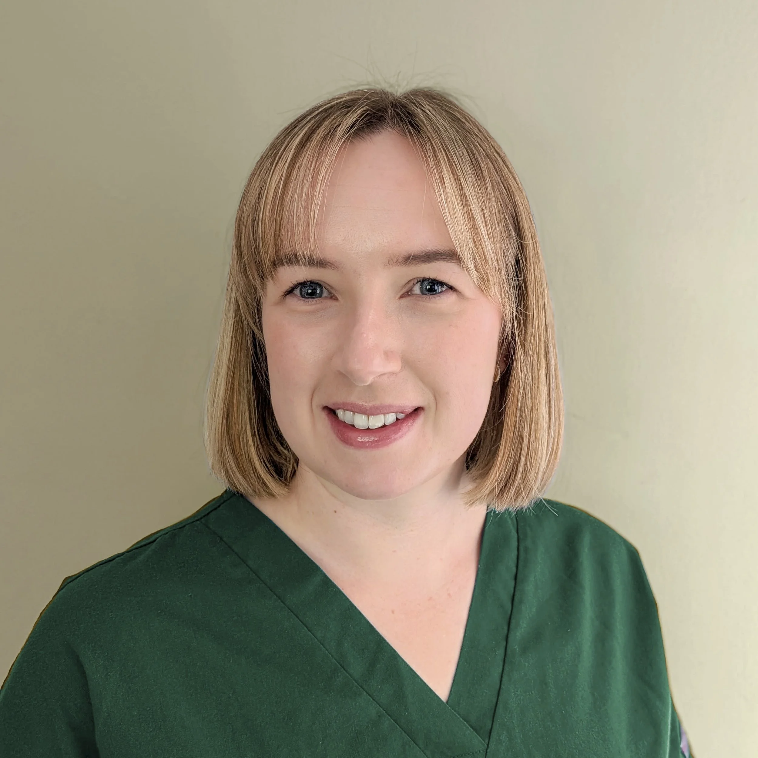 Portrait of a woman with shoulder-length blonde hair, wearing a green scrub top, smiling against a plain light-colored background.