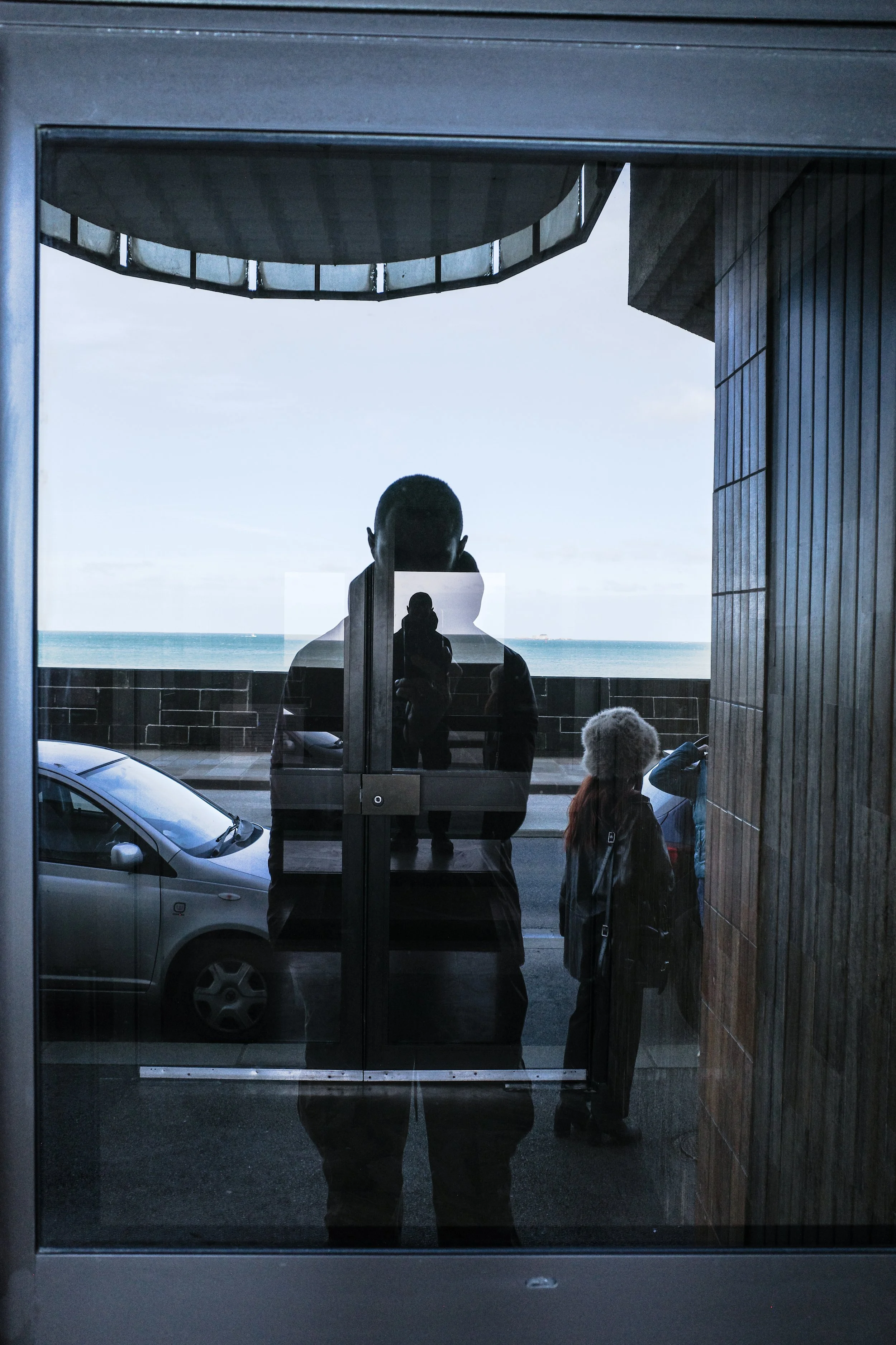 Reflected image of a person taking a photo outside a building with a view of the ocean, with other people and a parked car visible.