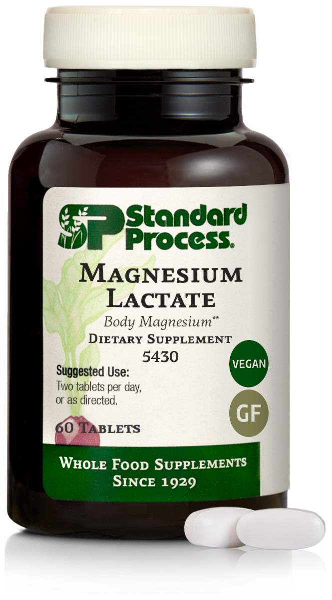 A bottle of Standard Process Magnesium Lactate dietary supplement with two white tablets in front, green and white label indicating vegan and gluten-free, and green band stating "Whole Food Supplements Since 1920."