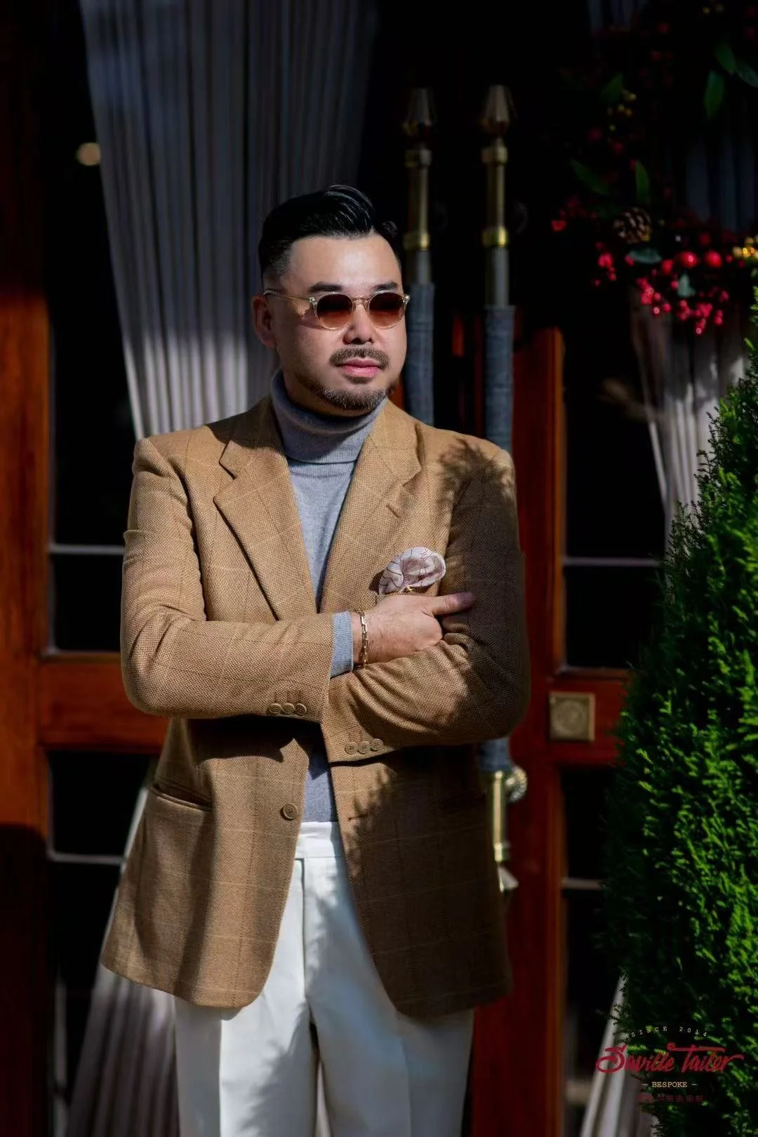A man wearing sunglasses, a beige blazer over a gray turtleneck, and white pants, standing outdoors with arms crossed and a serious expression.