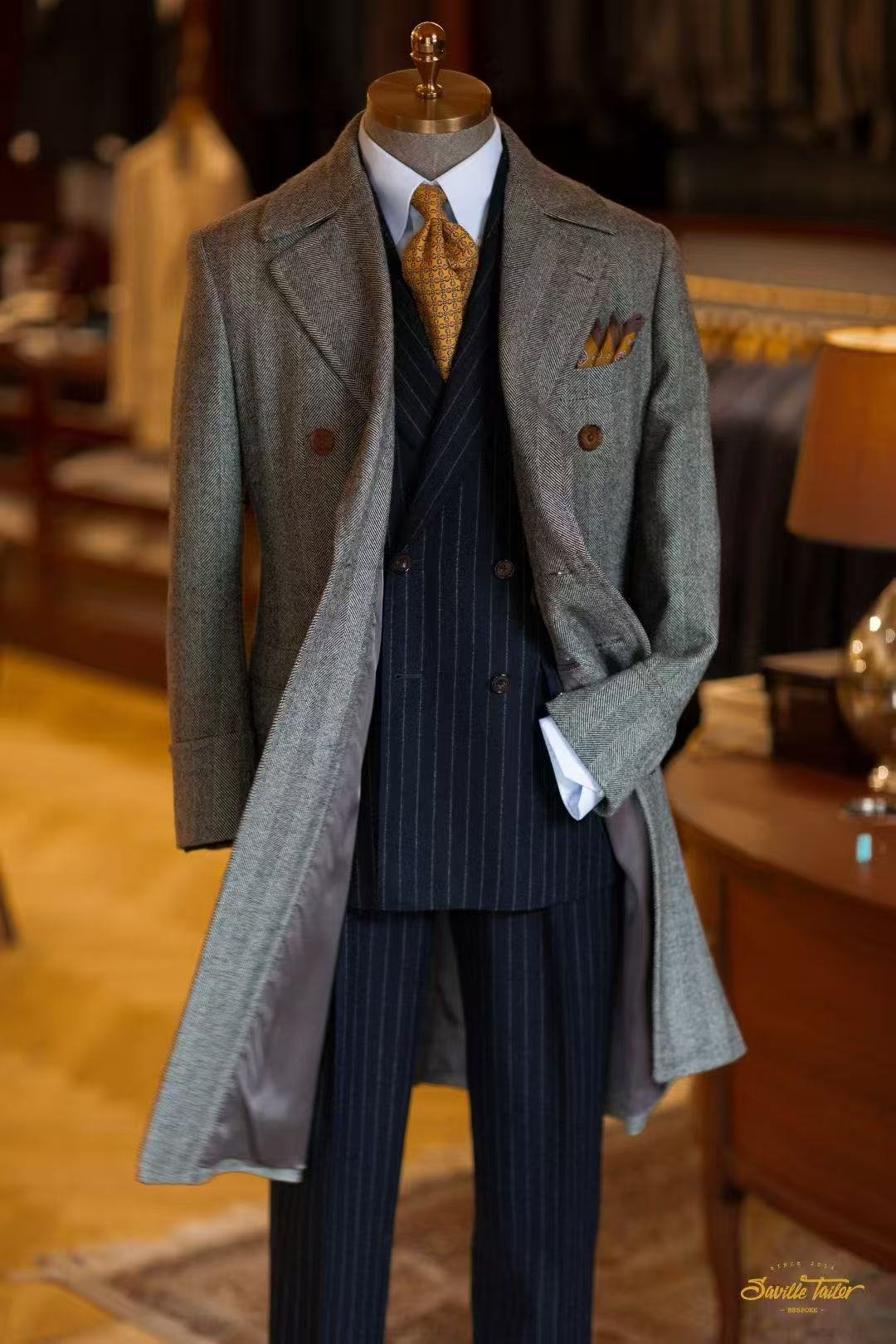 A men's mannequin dressed in a vintage style suit with a gray overcoat, pinstripe navy vest and pants, white dress shirt, and a patterned yellow tie, displayed in a clothing store or boutique.