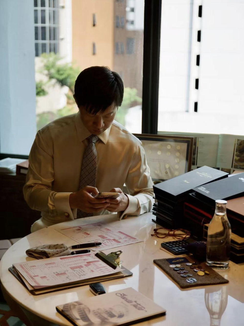 A man in a bespoke dress shirt and tie sitting at a cluttered desk in front of a large window, looking at his phone.