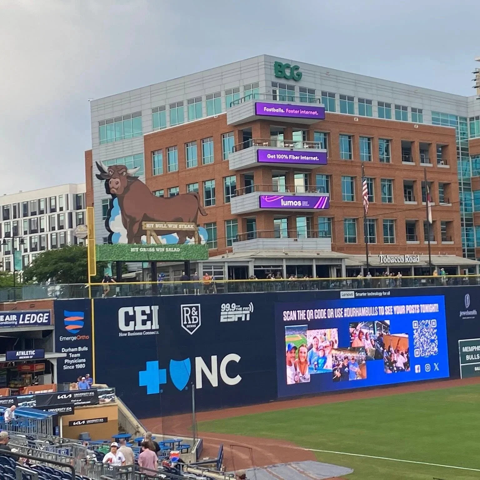 Billboard at a baseball stadium near Durham Bulls Athletic Park with digital ads, a large cartoon bull mascot holding a sign that reads 'Hit Bull Win Steak, Hit Greens Win Salad,' and a building with banners advertising internet services.