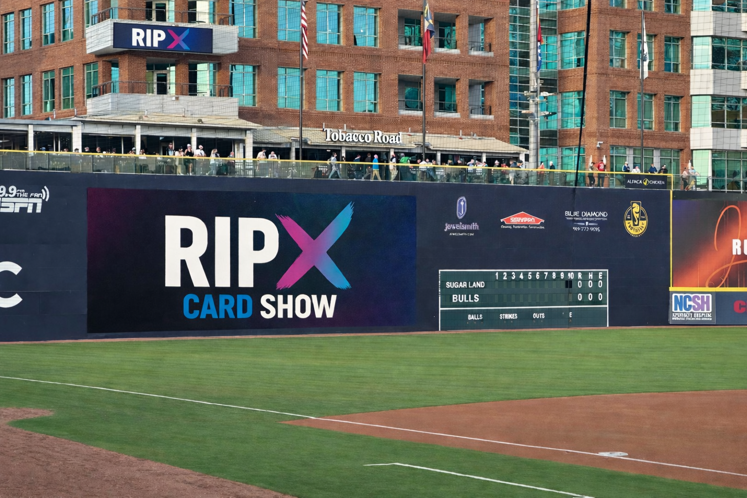 Baseball outfield wall with advertisements, scoreboard, and spectators above a grassy field in an urban stadium.