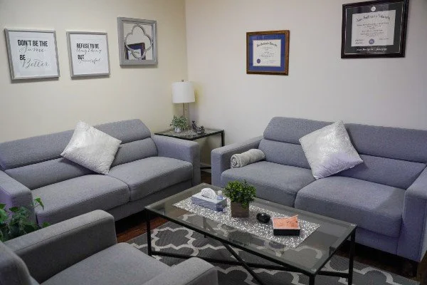 Systemic Solutions therapy room with two gray sofas and a matching armchair, decorated with silver pillows. A glass coffee table with a plant, tissues, and other items is in the center.
