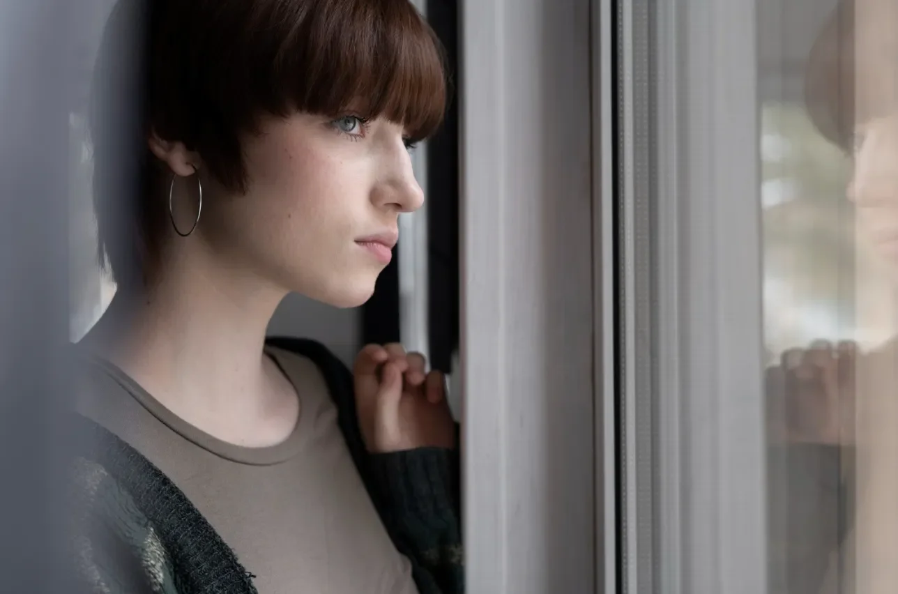 A woman with short red hair and hoop earrings looks out a window with a contemplative expression.