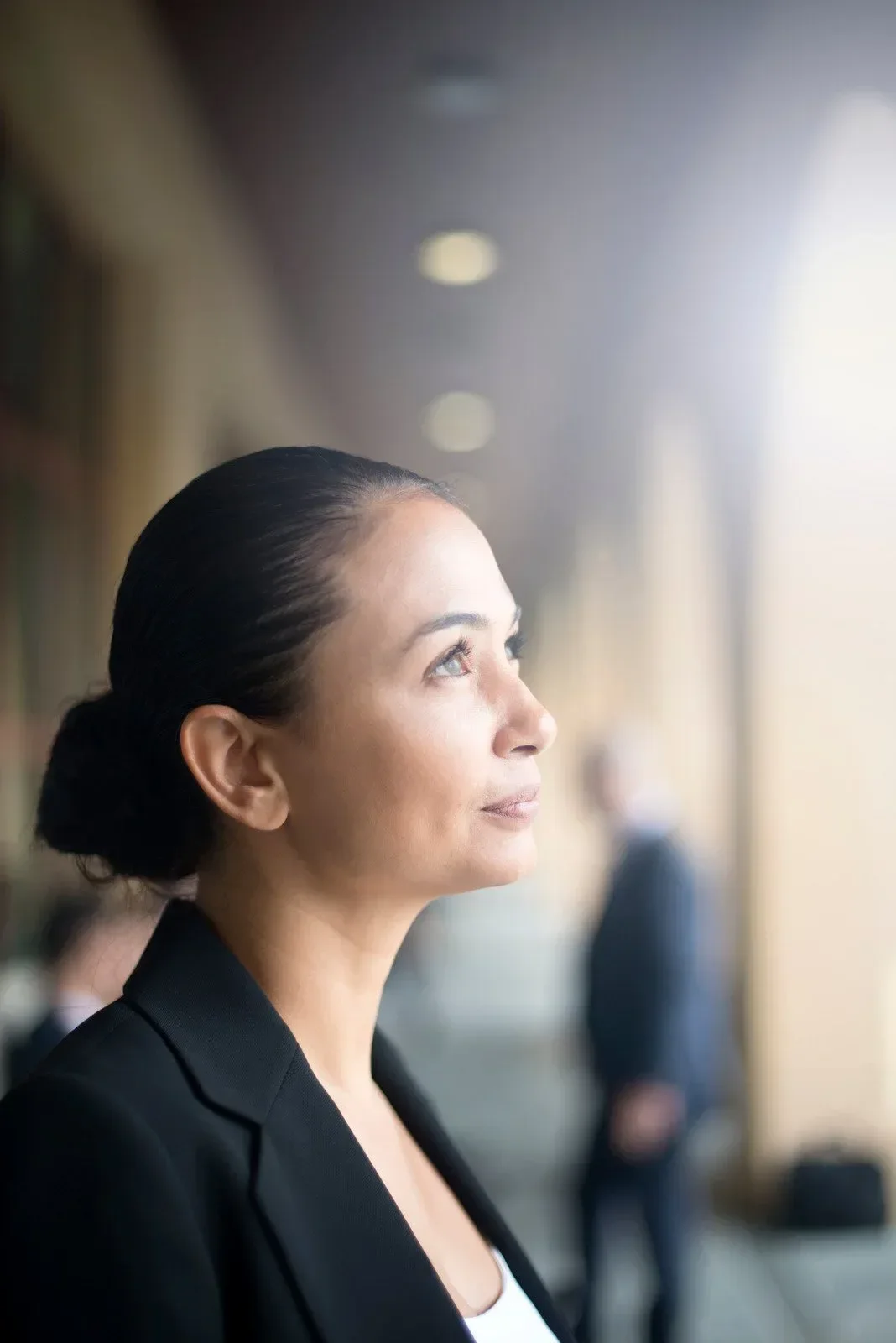 Side profile of a woman with dark hair in a bun, wearing a black blazer, looking thoughtfully out of a bright window.