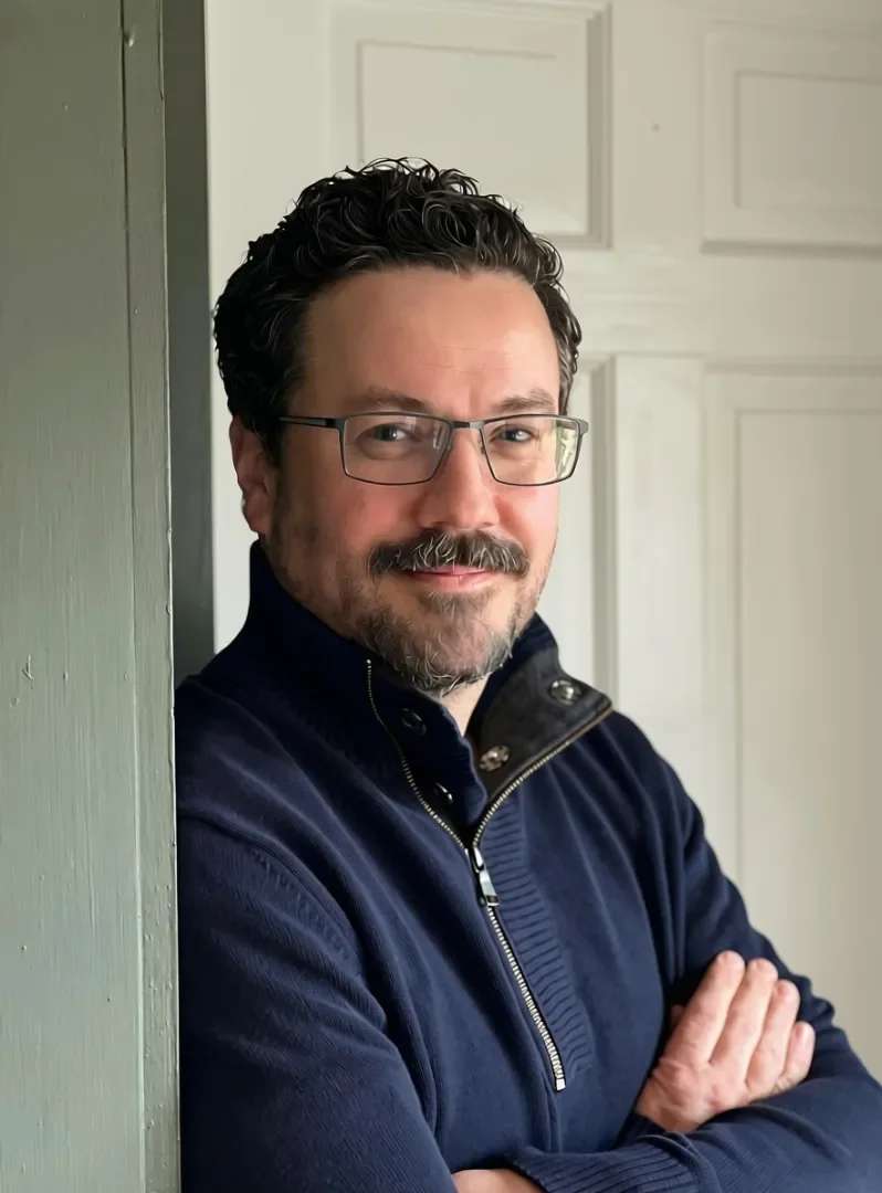 Dr. Christopher Rutt with dark, curly hair, glasses, and a beard, wearing a navy blue jacket with a zipper, standing indoors with arms crossed, smiling at the camera.