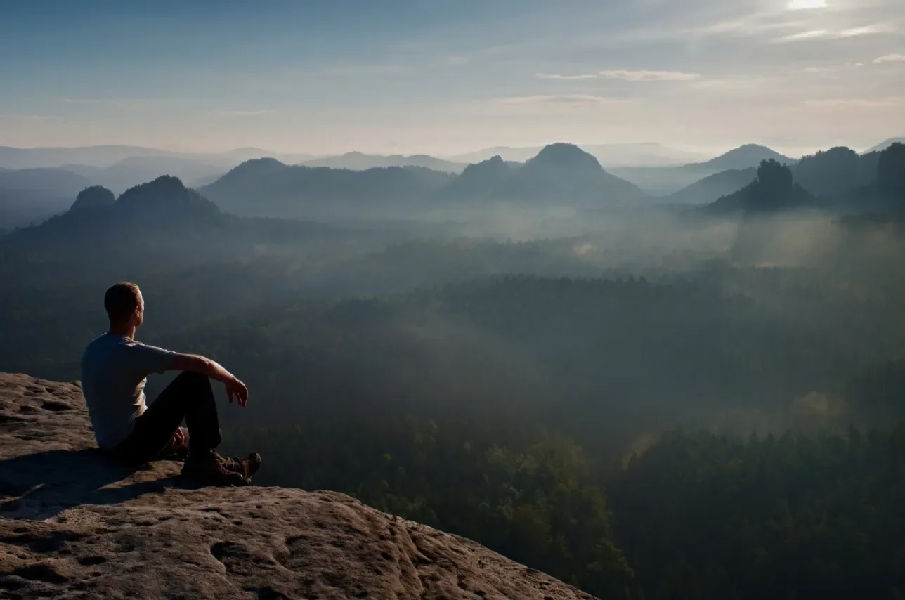A person sitting on a rocky ledge overlooking a foggy mountain landscape during sunrise or sunset.