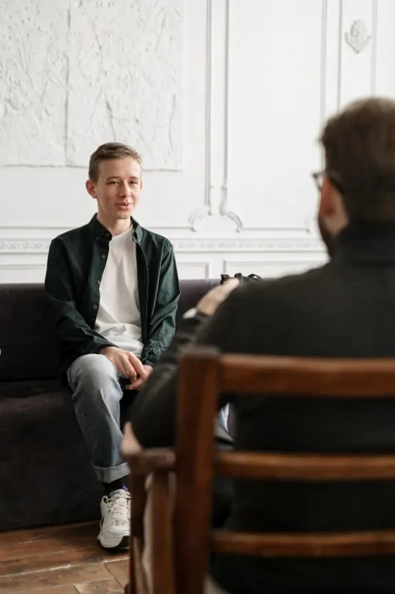 A young man sitting on a black couch being interviewed by an older man in a black jacket, with a white wall and decorative molding in the background.
