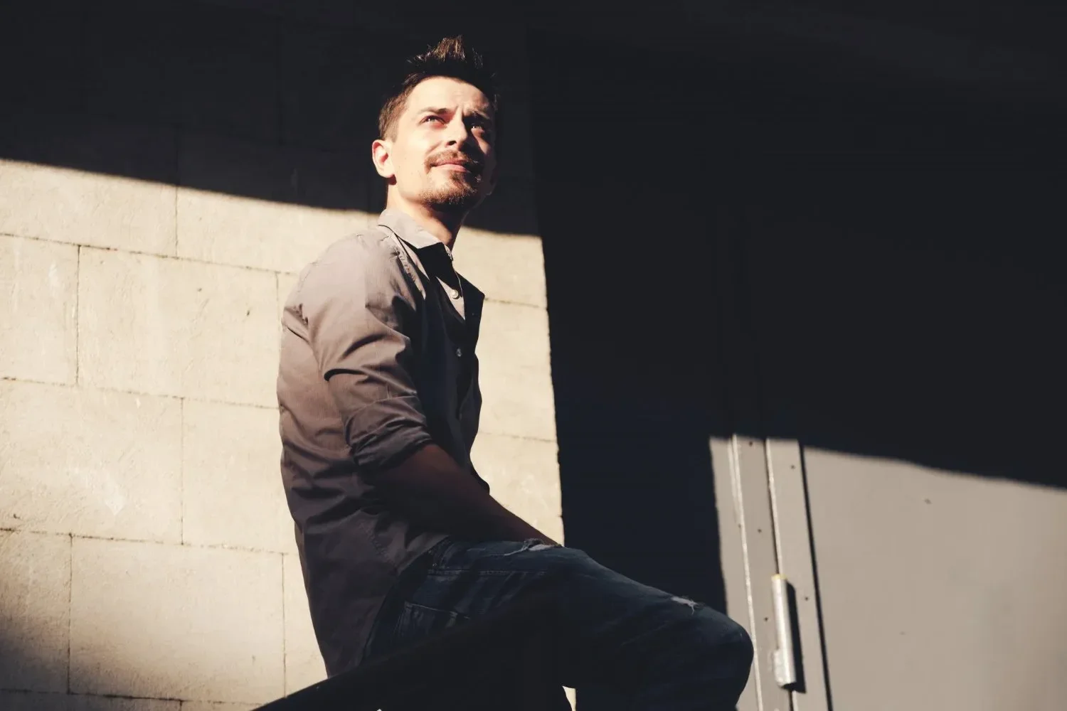 A man with short brown hair and a goatee sitting against a concrete wall, looking slightly upwards with sunlight on his face, wearing a gray shirt and jeans.