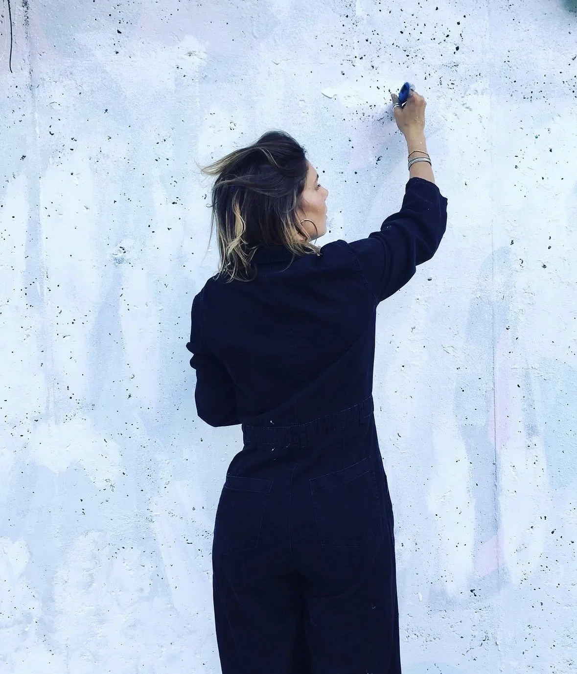 A woman writing on a white wall with black specks, using a blue chalk or marker, facing the wall with her back to the camera, wearing a black jacket and high-waisted jeans.