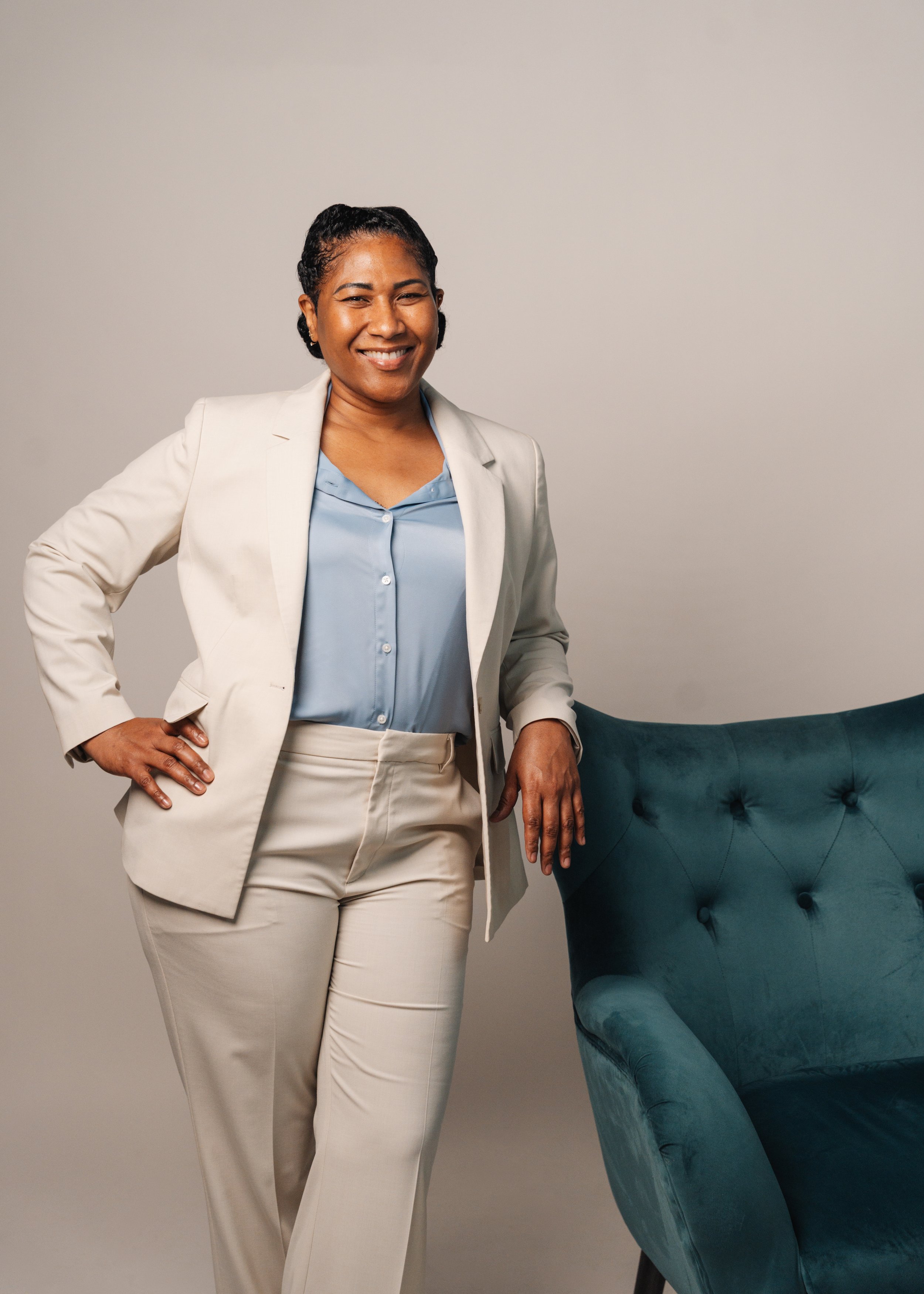 A woman with short curly black hair, wearing a light beige suit with a matching pair of pants and a pale blue button-up shirt, standing with her hand on her hip next to a blue tufted chair in a neutral-colored room.