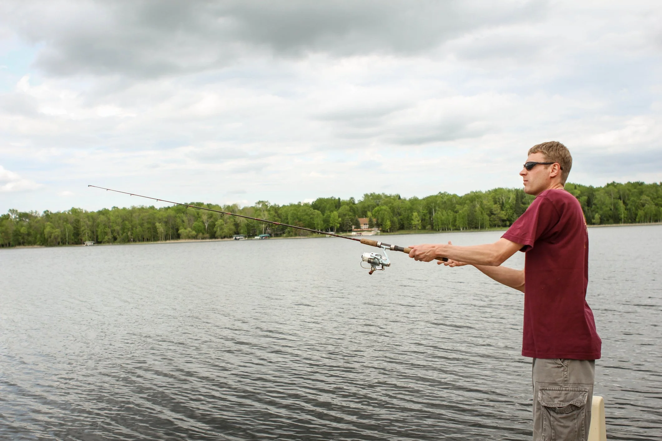 A man wearing sunglasses and a maroon T-shirt is fishing in a lake, standing near the shore with a fishing rod in his hands. The background features a cloudy sky, a treeline, and some houses across the water.