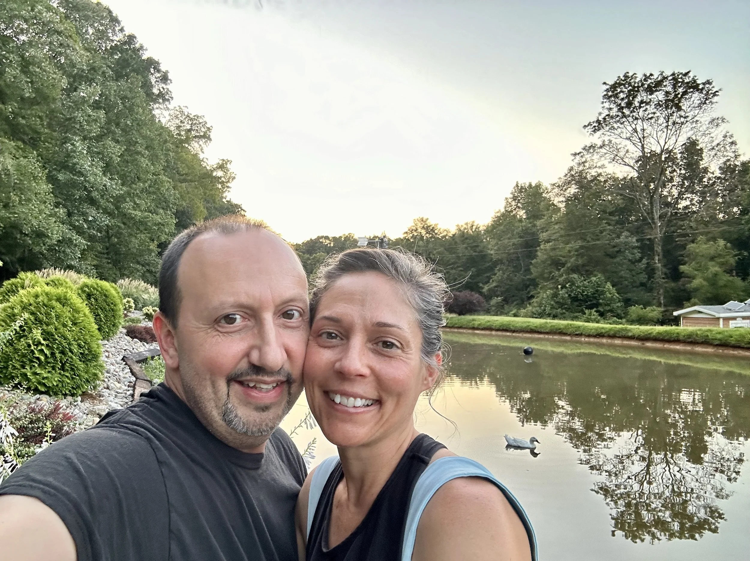 A smiling middle-aged man and woman taking a selfie outdoors near a pond with ducks, surrounded by lush green trees and plants, during what appears to be late afternoon or early evening.