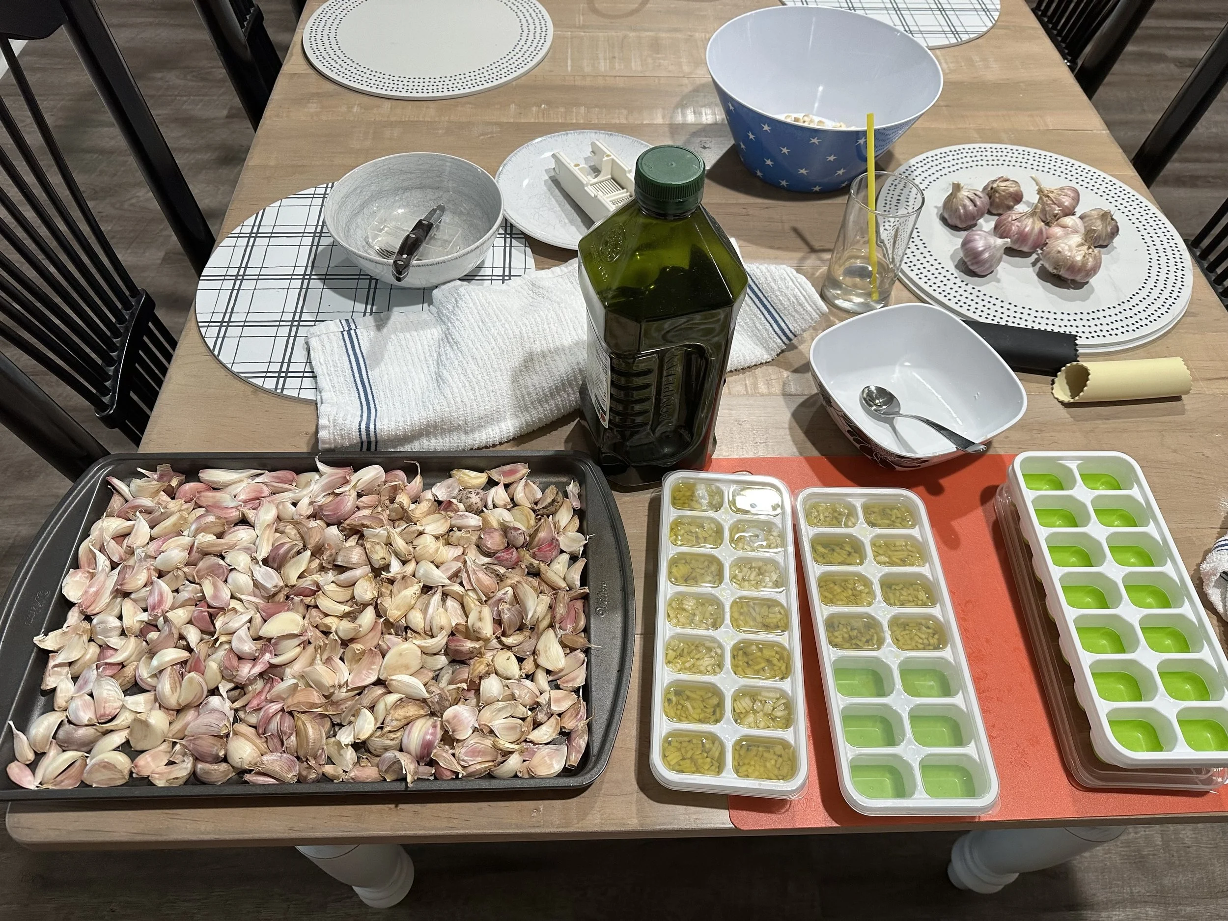 A wooden dining table with various cooking ingredients and kitchen utensils, including a large tray of garlic cloves, a bottle of olive oil, garlic bulbs on a plate, small containers of chopped garlic, molds for making garlic cubes, empty bowls, and a glass with a yellow straw.