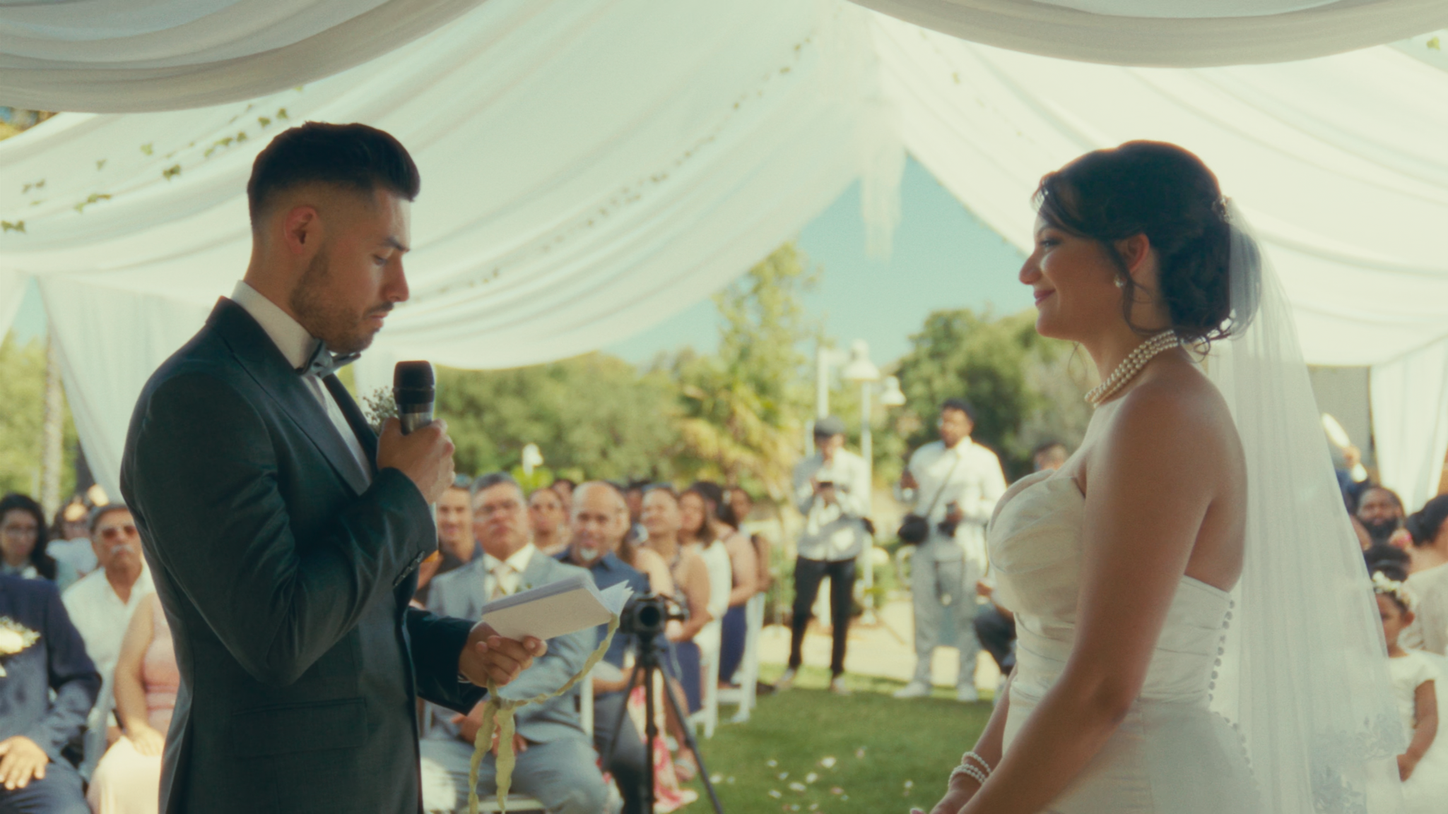 A groom in a black tuxedo reading vows from a notebook during a wedding ceremony, while the bride in a strapless wedding dress and pearl necklace listens with a smile, under a white tent with outdoor guests in the background.