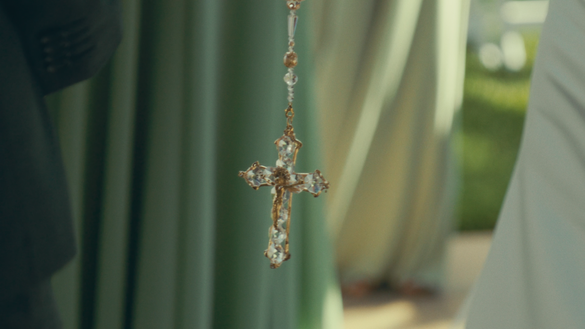 A close-up of a decorative cross pendant with embedded gemstones hanging from a chain, set against a blurred background of green and white curtains.