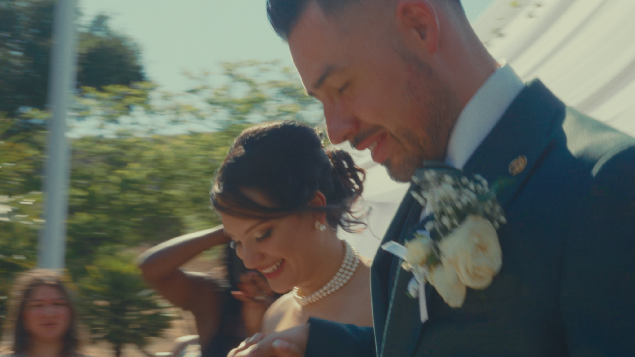 A smiling man in a suit with a boutonniere and a woman with jewelry, both looking happy, during a wedding celebration outdoors on a sunny day.